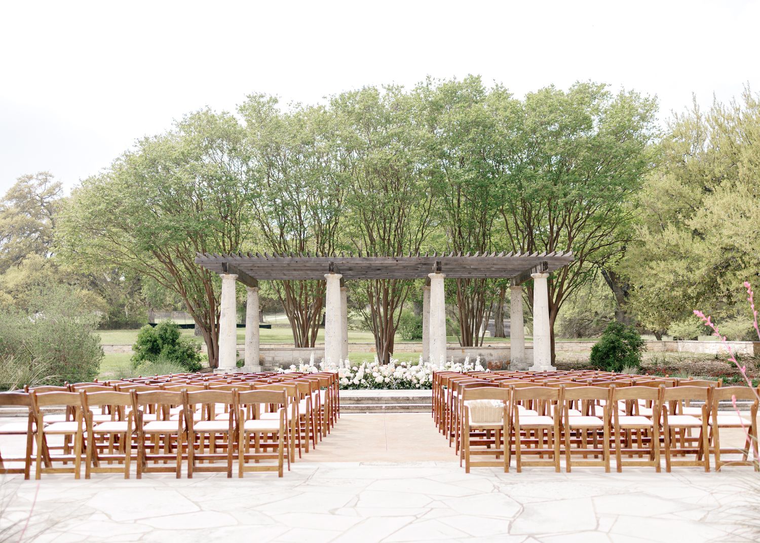 An elegant outdoor wedding ceremony setup with rows of wooden chairs facing a white-draped pergola amid lush green trees.