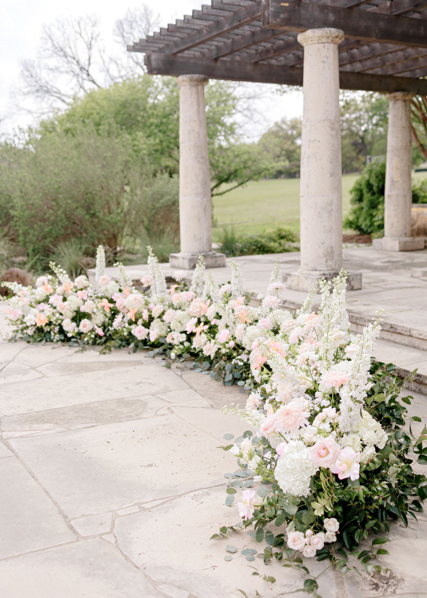 Elegant floral arrangement of pink and white flowers lines a stone pathway under classical columns with a pergola.
