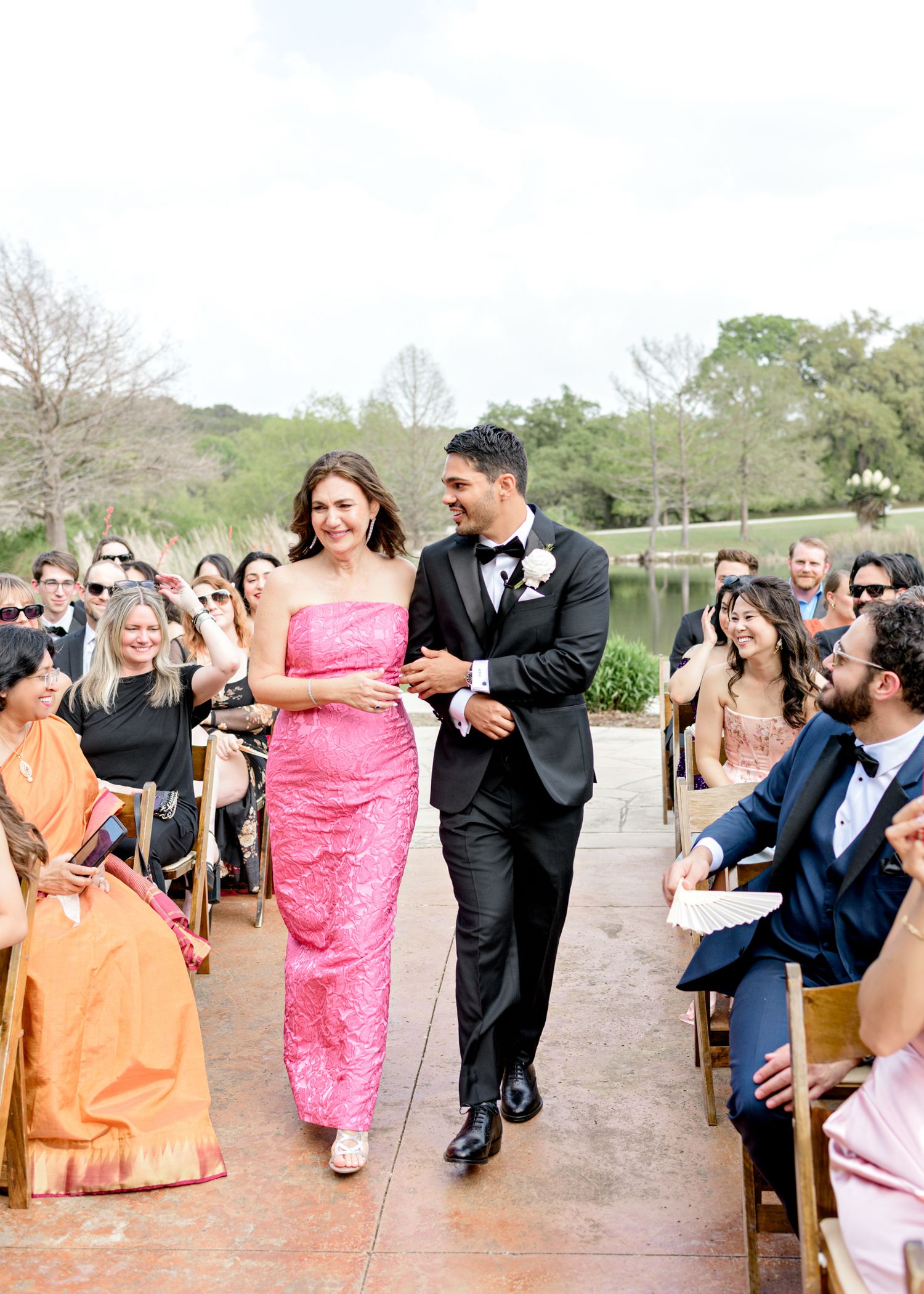 A couple with their wedding party walk down an outdoor aisle as wedding guests smile and cheer from their seats.