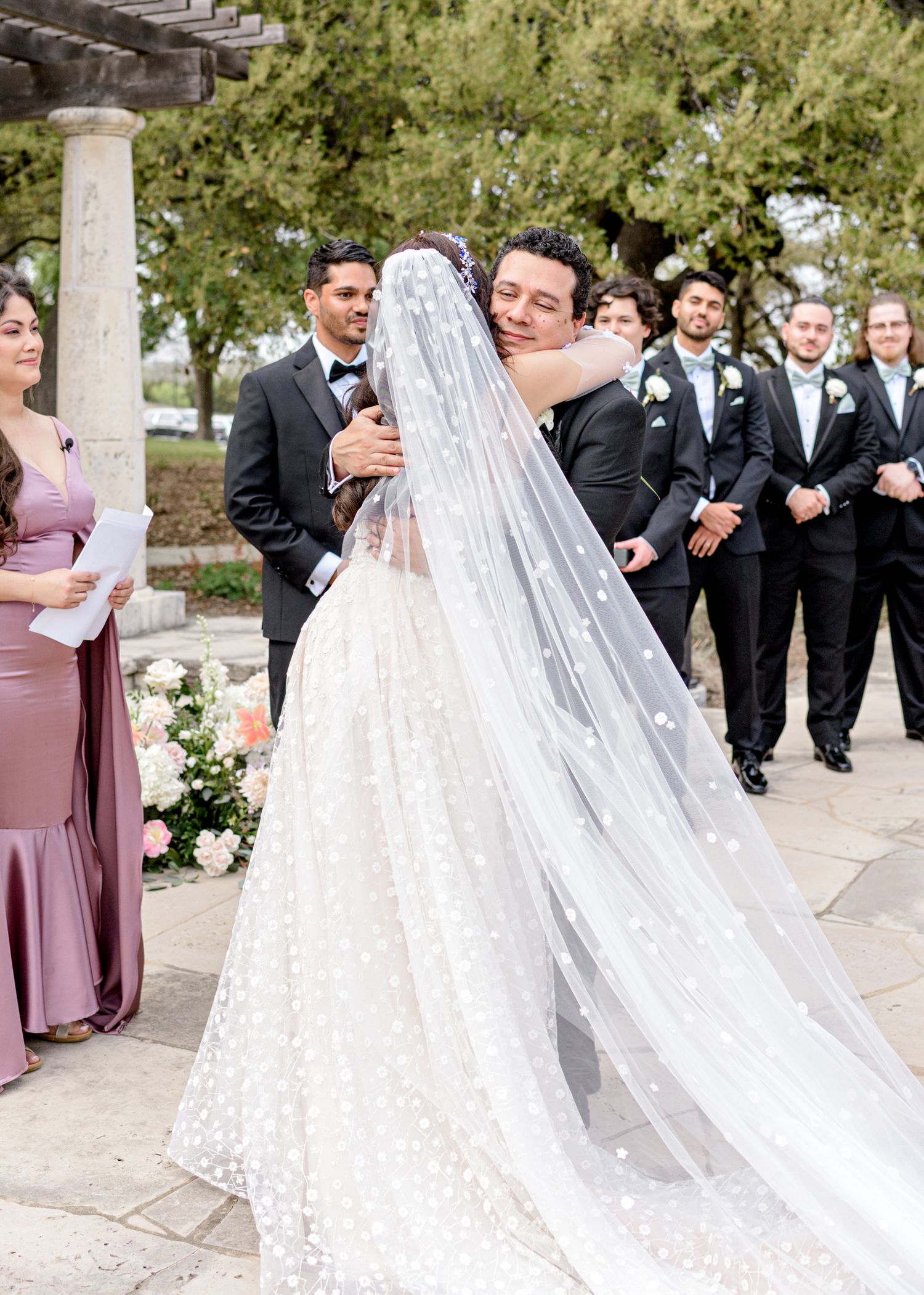 The newlywed couple embraces during their wedding ceremony while the bridal party looks on at an outdoor venue.