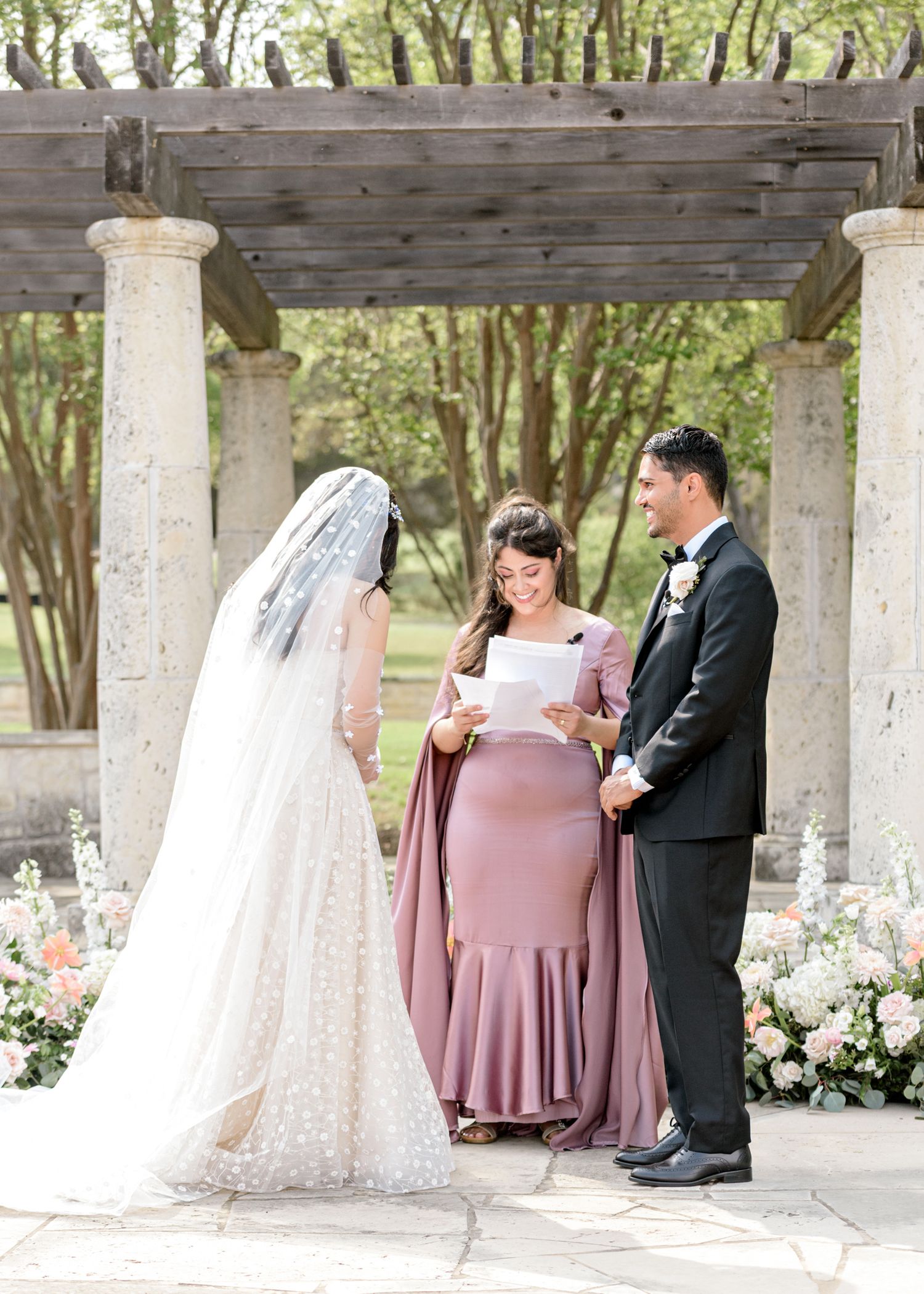 A romantic outdoor wedding ceremony takes place under a rustic wooden pergola with white stone columns and floral decorations.