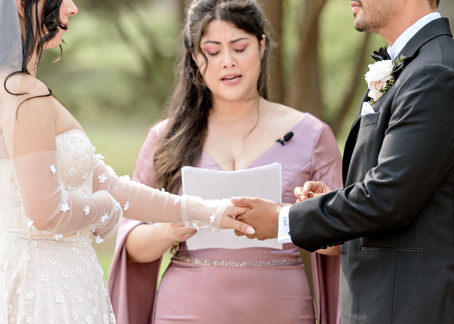 A wedding officiant in a mauve dress reads from papers during an outdoor ceremony in a garden setting.