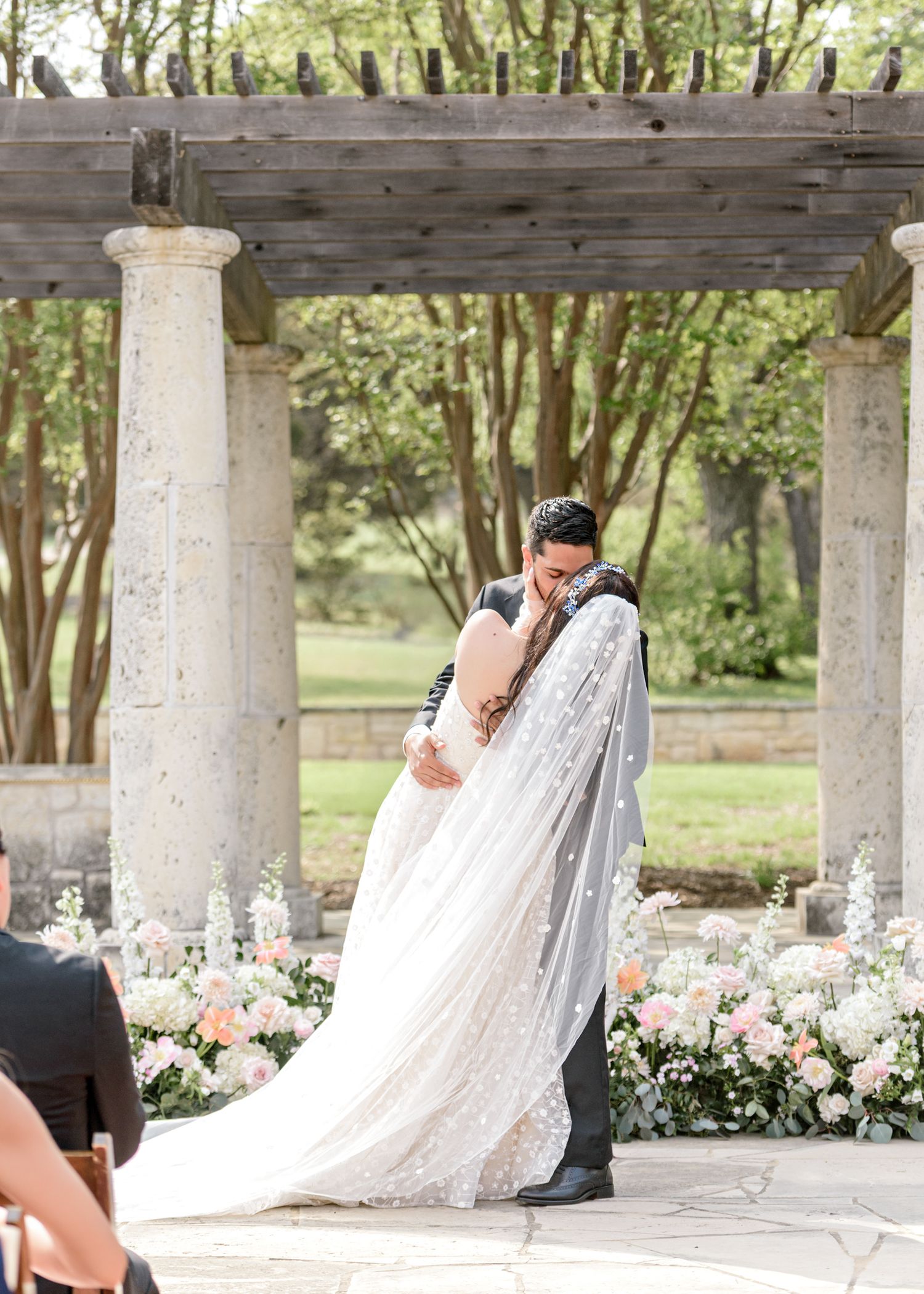 A romantic moment during a wedding ceremony beneath a columned pergola surrounded by pink floral arrangements.