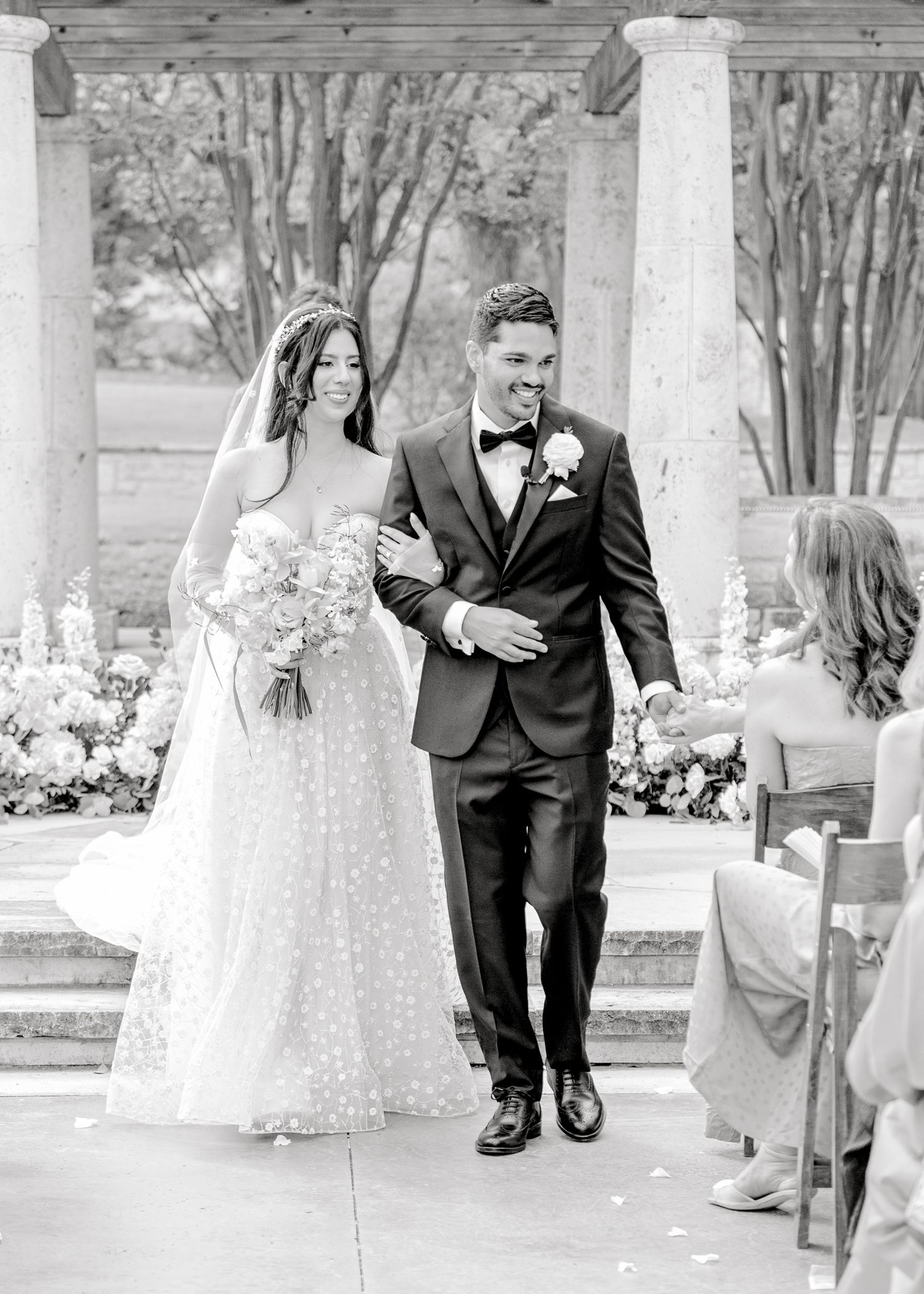 Black and white photograph of a couple on their wedding day walking arm in arm along an outdoor garden path.