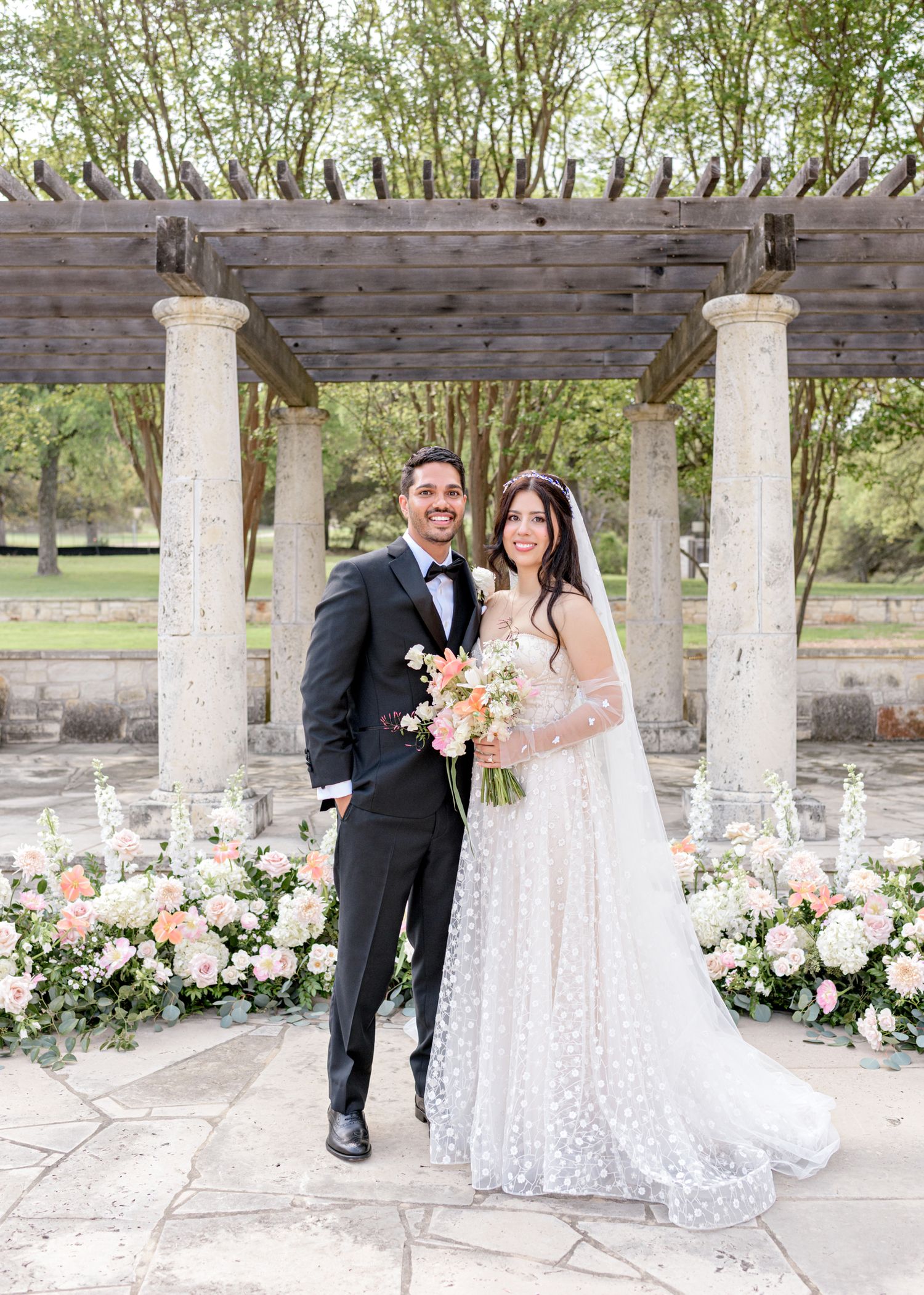 Newlyweds pose under a rustic wooden pergola with stone columns surrounded by pink and white floral arrangements.