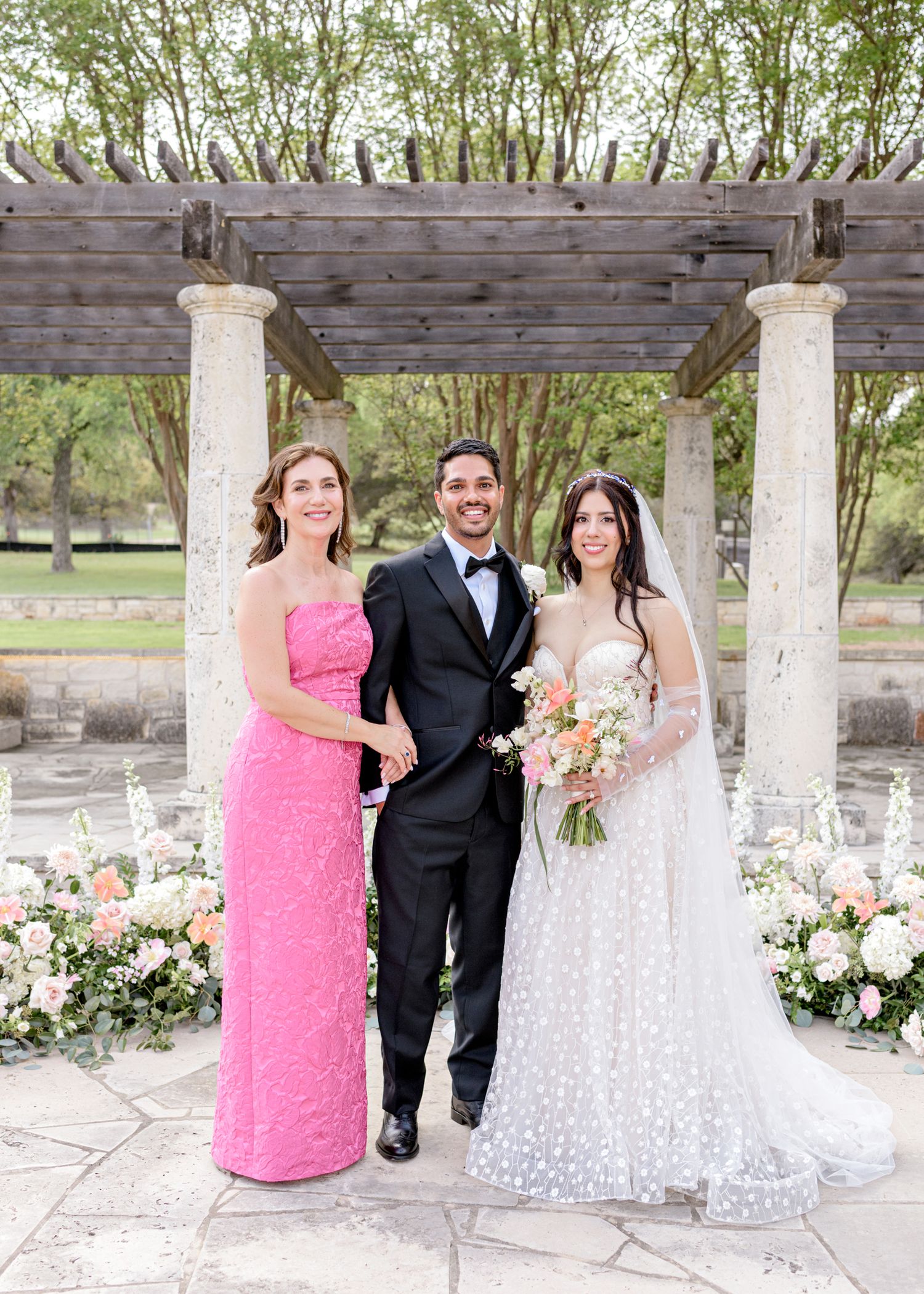 A wedding party poses under a stone pergola decorated with pink and white floral arrangements on a sunny day.