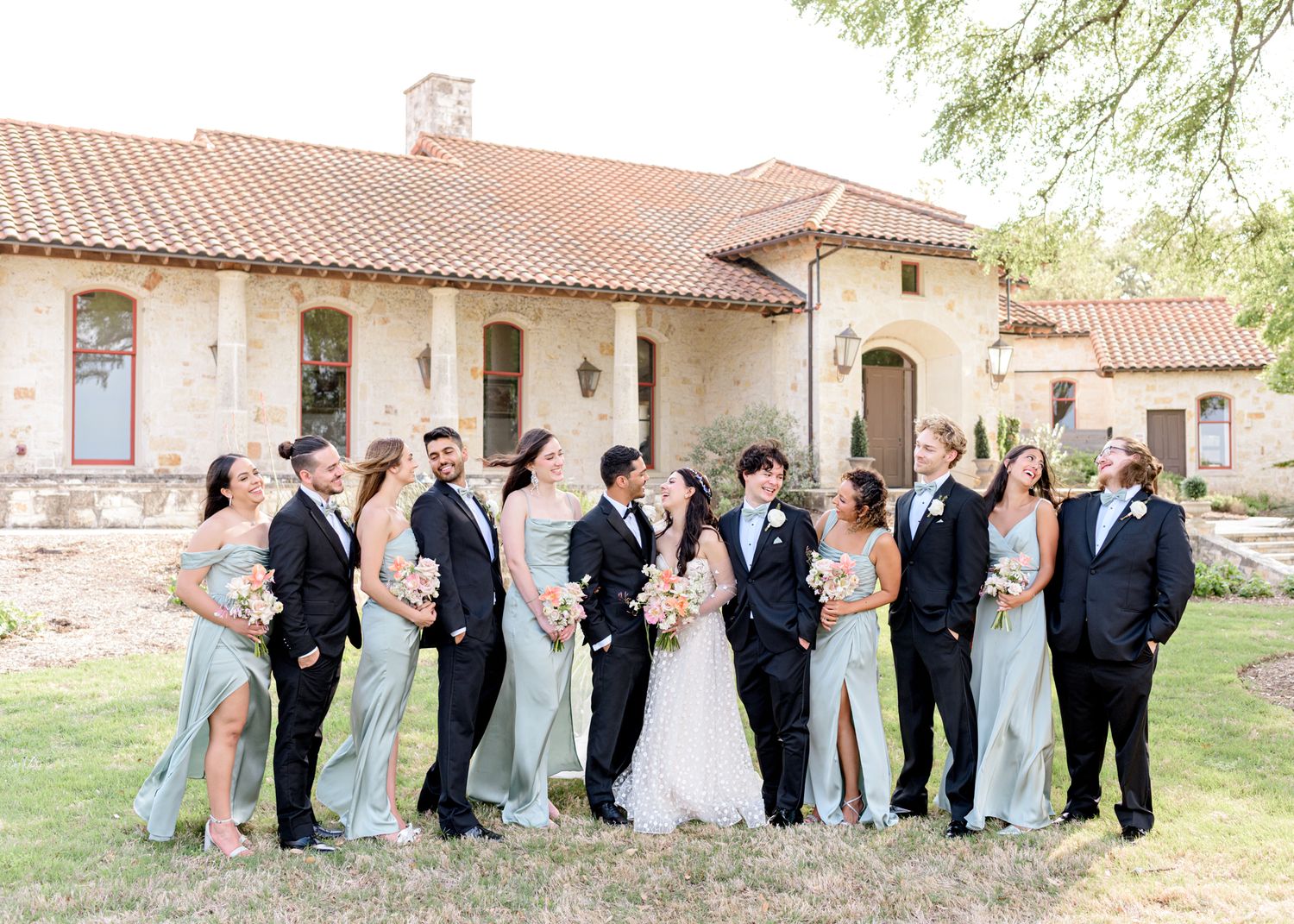 A bridal party wearing light blue and black formal attire poses together on a lawn in front of a Spanish-style building.