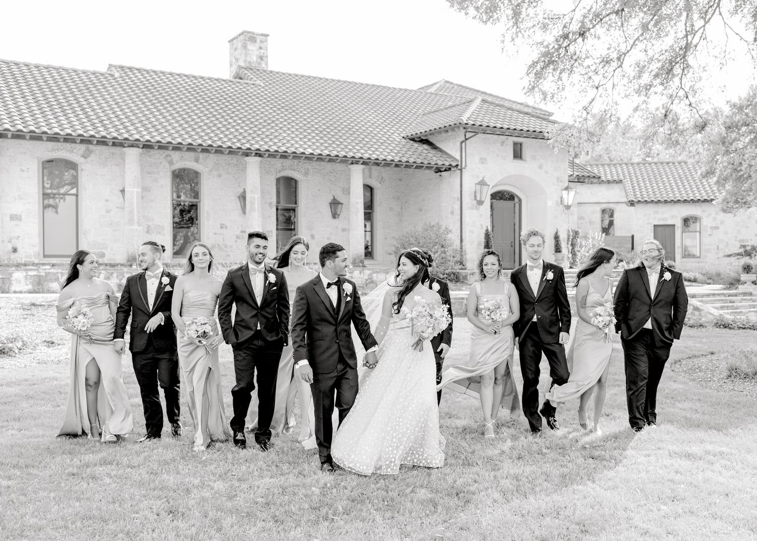 Wedding party walks together outside a Mediterranean-style building in black and white photography.