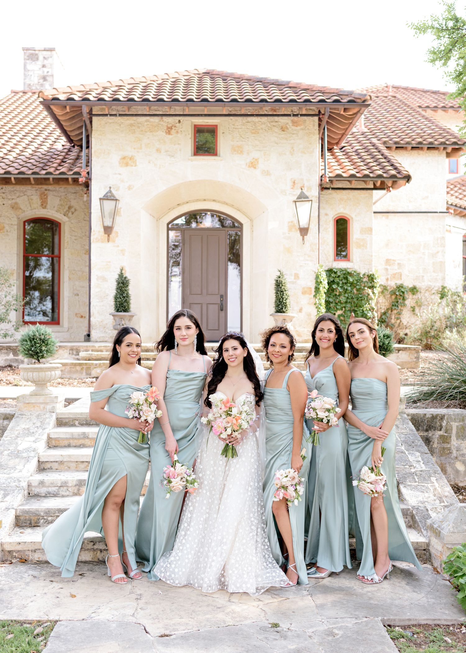 Bridal party in sage green dresses poses on stone steps in front of Mediterranean style villa entrance.