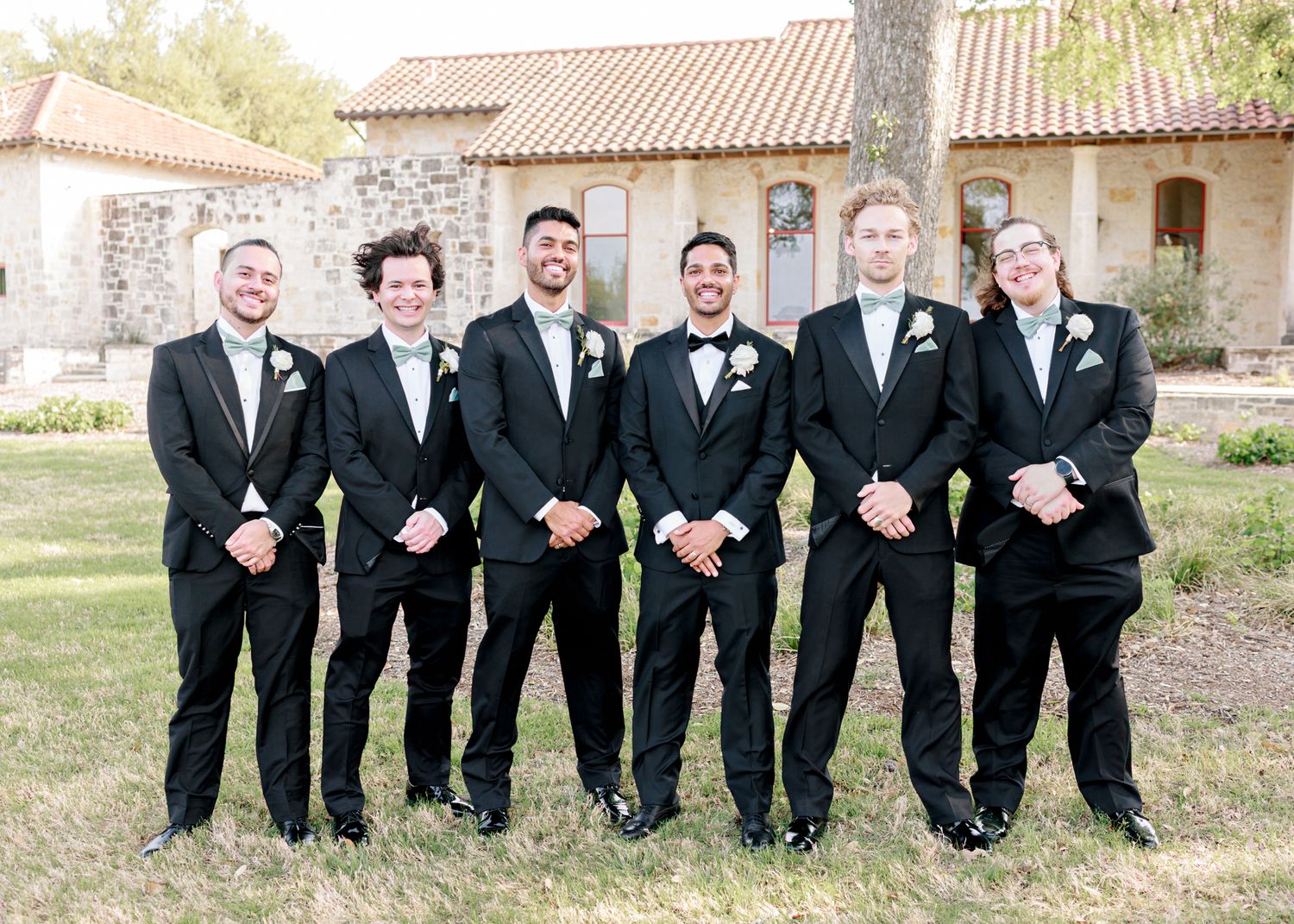 Groomsmen in black tuxedos stand in line smiling outside Spanish-style building.