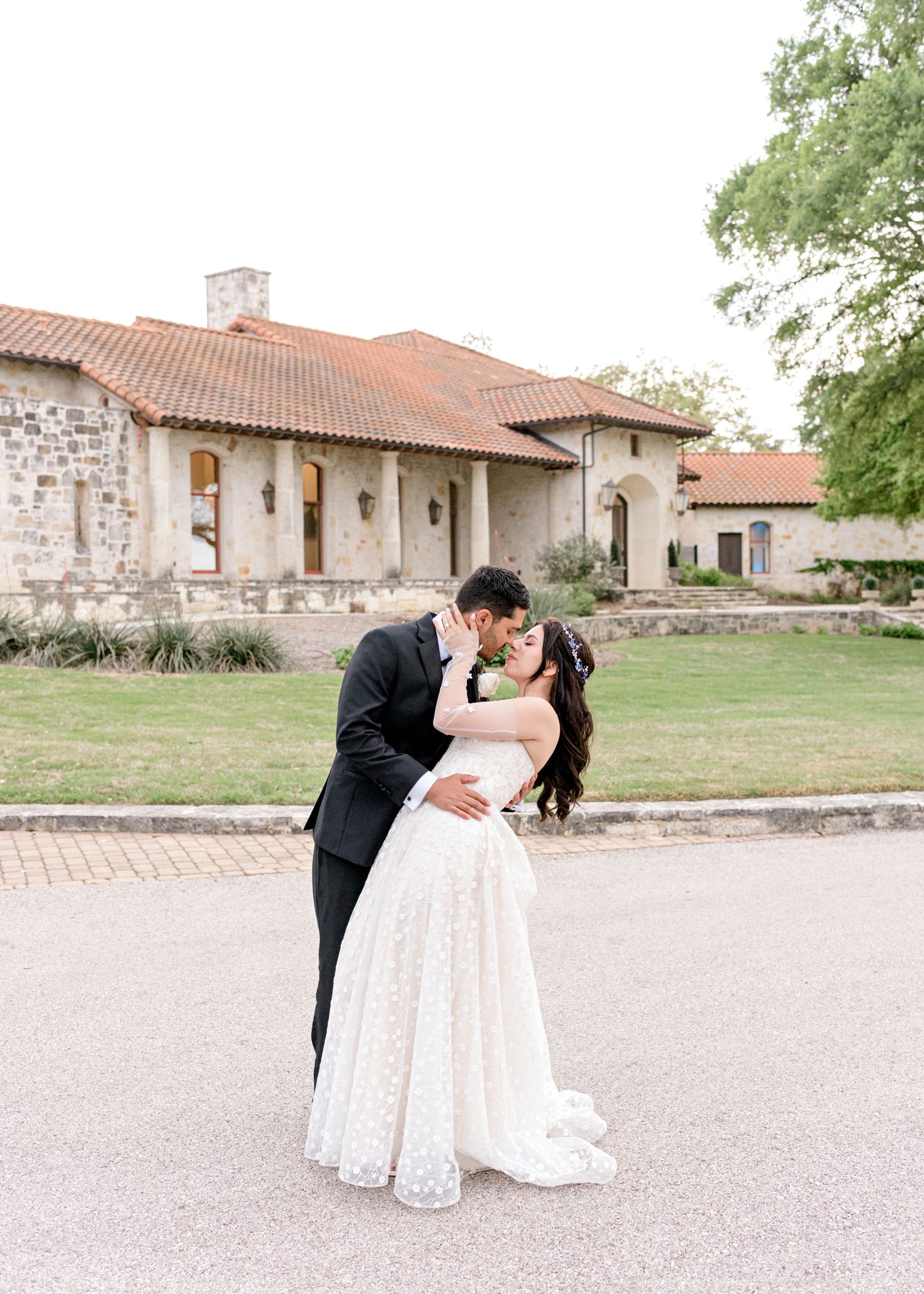 A romantic wedding couple share a dip-style kiss outside a rustic stone building with terracotta roof tiles.