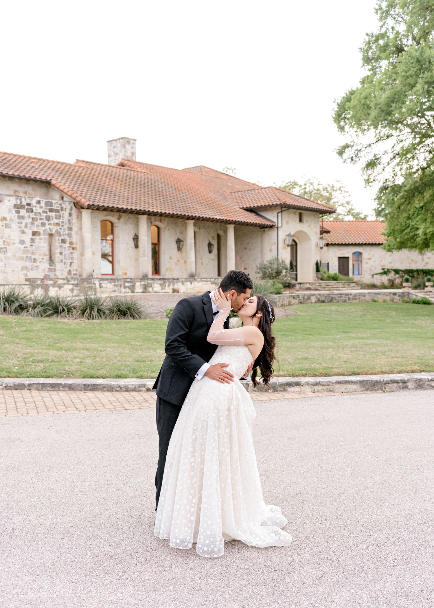 A romantic wedding couple embraces in front of a rustic stone building with terra cotta roof tiles.