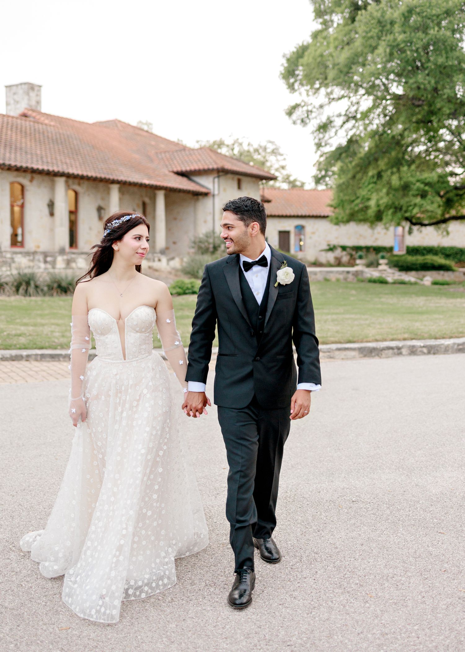 A bride in a white lace gown and groom in black tuxedo walk together on a path outside a Spanish-style building.