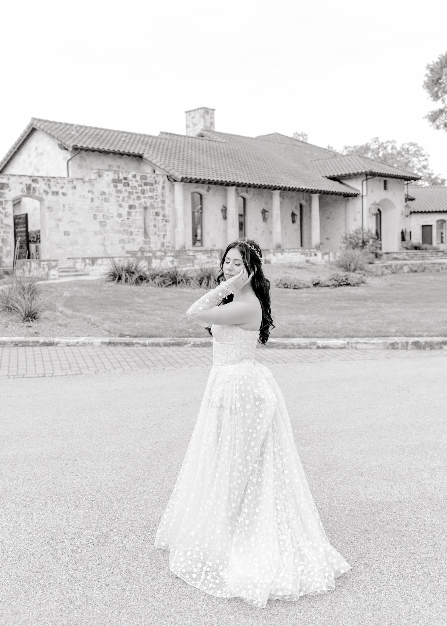 A black and white photograph shows someone in a flowing lace wedding dress twirling on a paved pathway near a stone building.