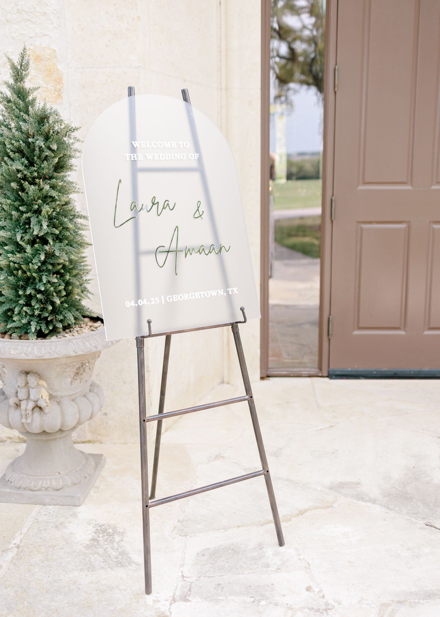 A clear acrylic welcome sign on a metal easel stand sits at an elegant entrance with a potted evergreen tree.