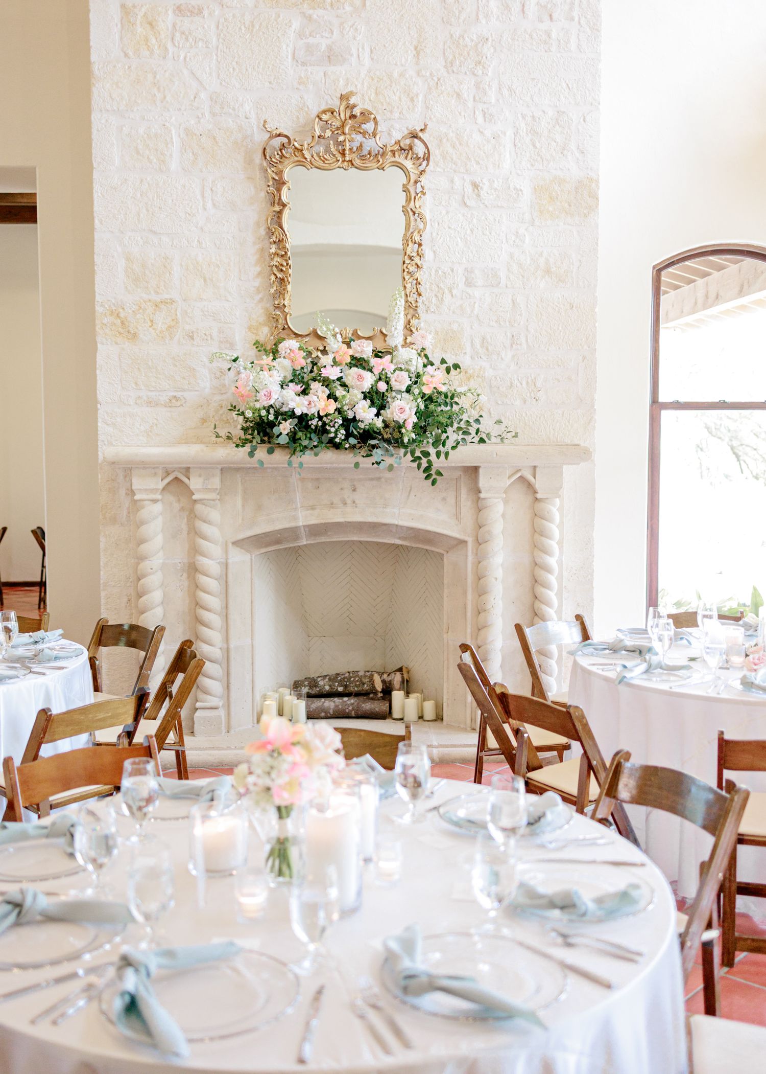 Elegant wedding reception setup with round tables, white linens, and ornate gilded mirror above a limestone fireplace.