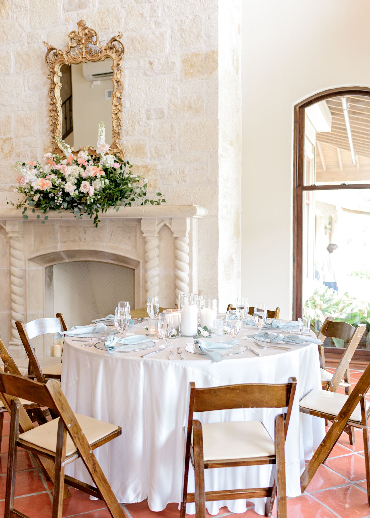 An elegant wedding reception table with white linens and wooden chairs beneath an ornate gold mirror and floral arrangement.