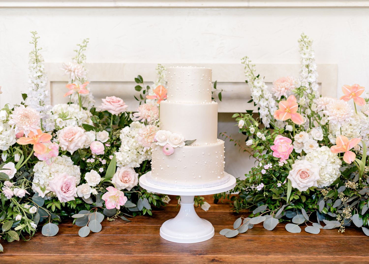 Elegant white wedding cake on pedestal with lush pink and white floral arrangements and eucalyptus greenery backdrop.