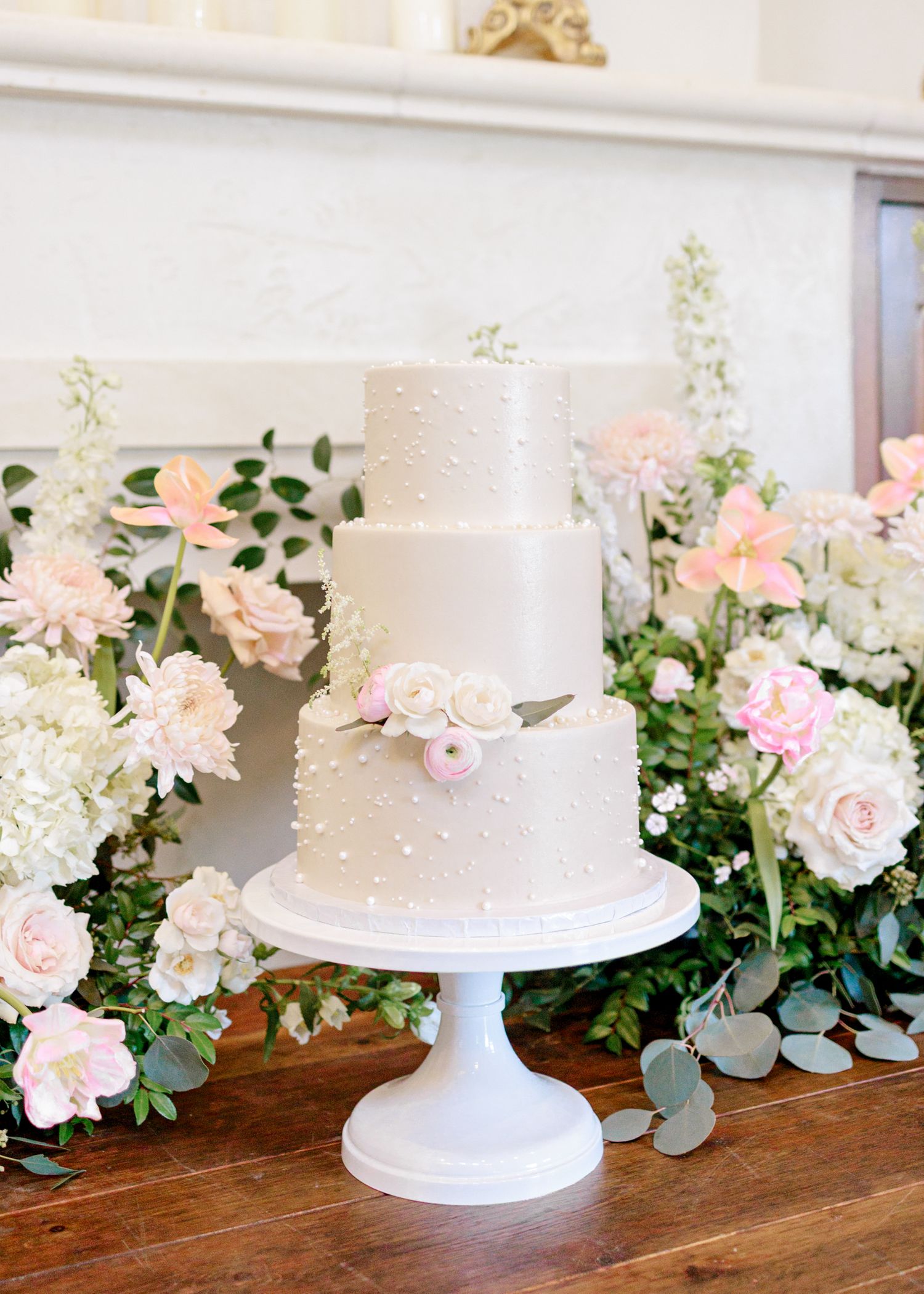 Elegant three-tiered white wedding cake with delicate pearl details and blush roses on display with floral arrangements.