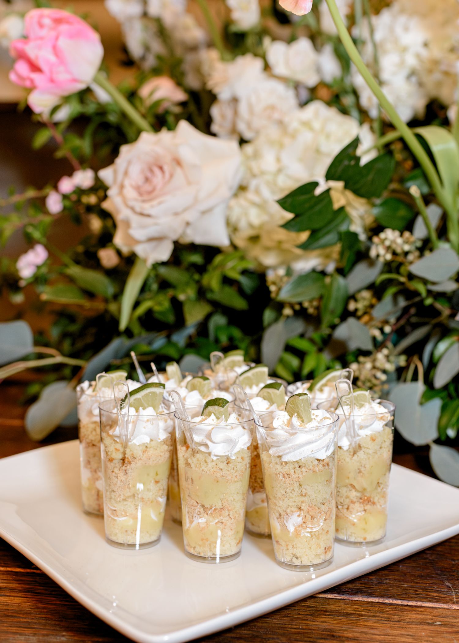 Mini dessert shooters with whipped cream garnish displayed on white plate beside pink and white floral centerpiece.