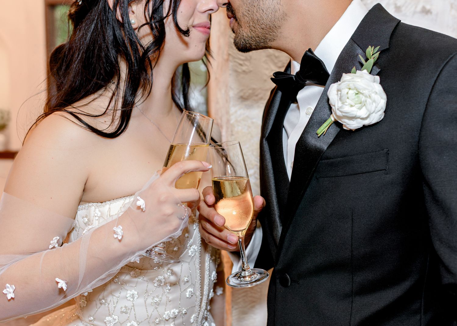 A close-up shot of newlyweds sharing champagne together with groom wearing black tuxedo with white boutonniere.