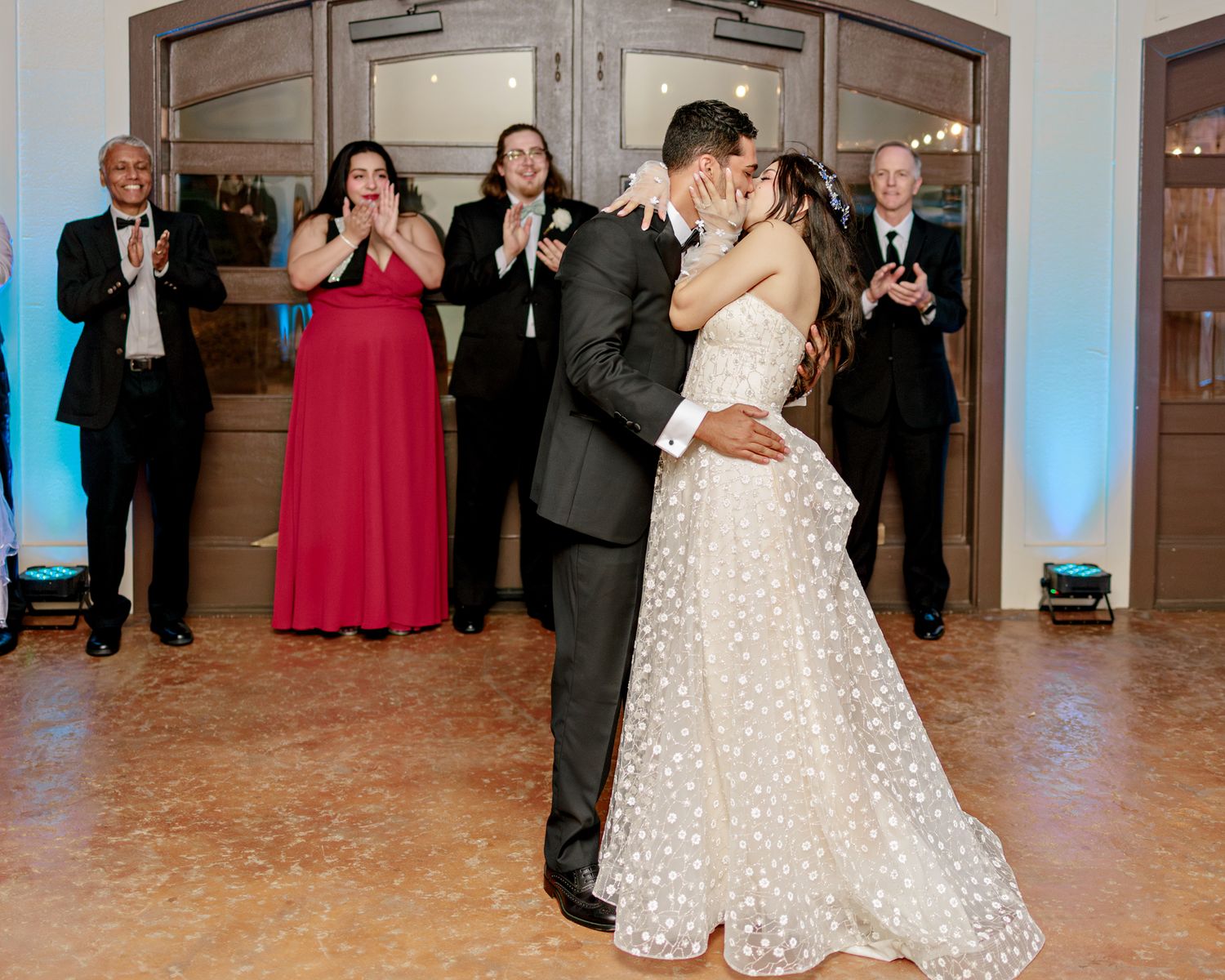 A couple shares their first dance at a wedding reception while guests in formal attire applaud in the background.