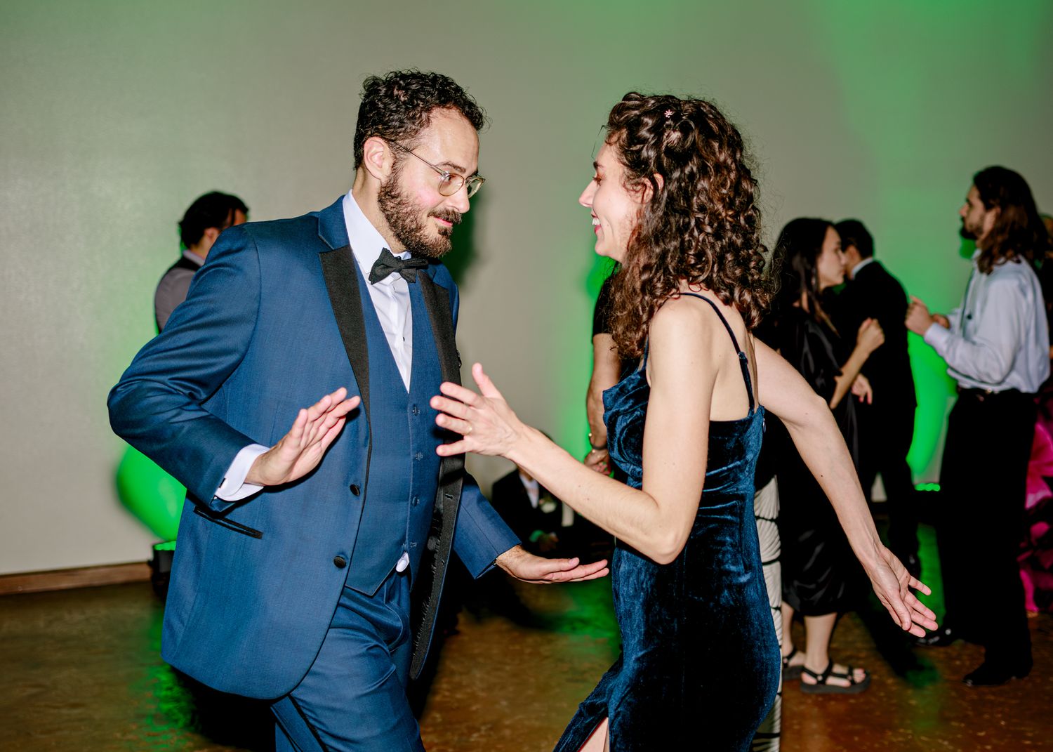 Two wedding guests share an energetic dance moment under green lighting at a reception.