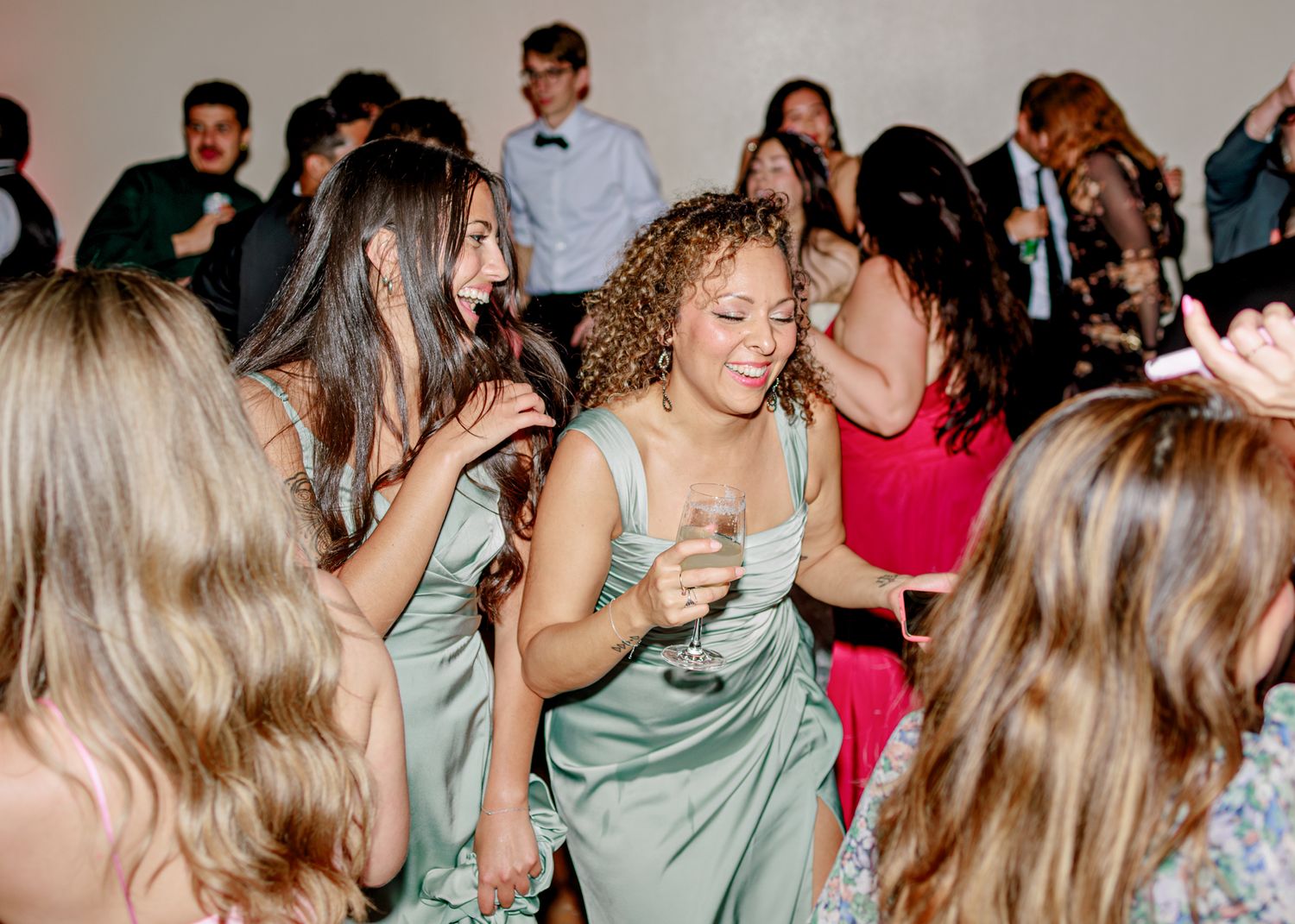 A group of people dancing and having fun at a lively party with some wearing sage green and red dresses.