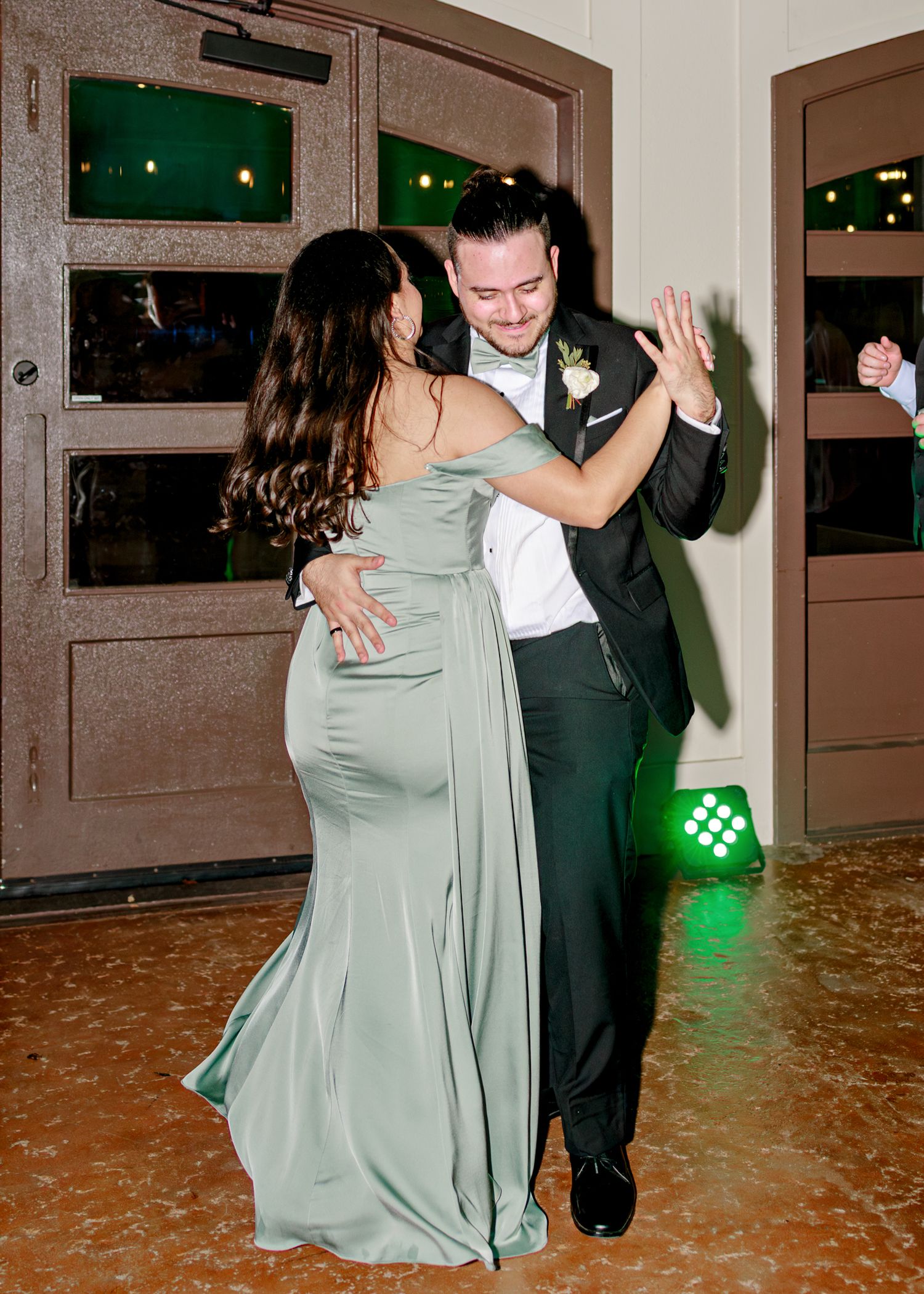A couple dances together at an elegant event, with the woman wearing a flowing sage green gown and man in formal attire.