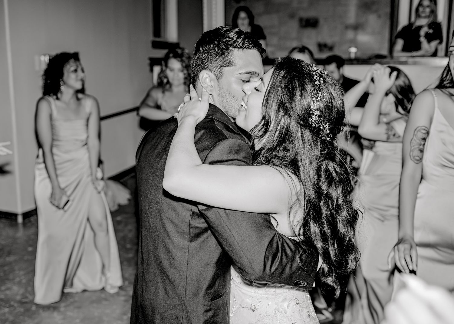 A couple shares their first dance at a wedding reception while guests look on in black and white.