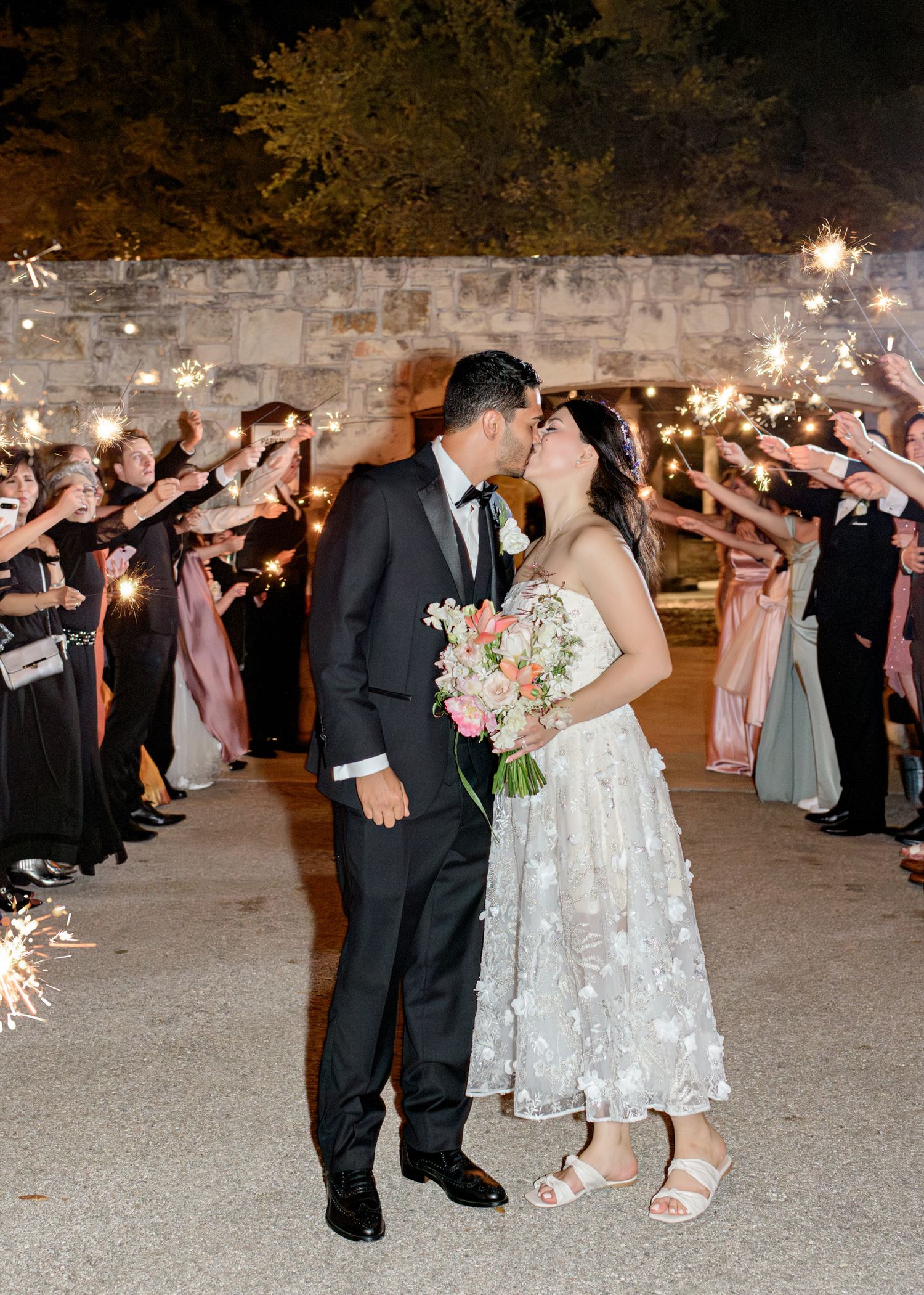 Newlyweds share a romantic kiss during sparkler sendoff with wedding guests lining pathway in evening celebration.