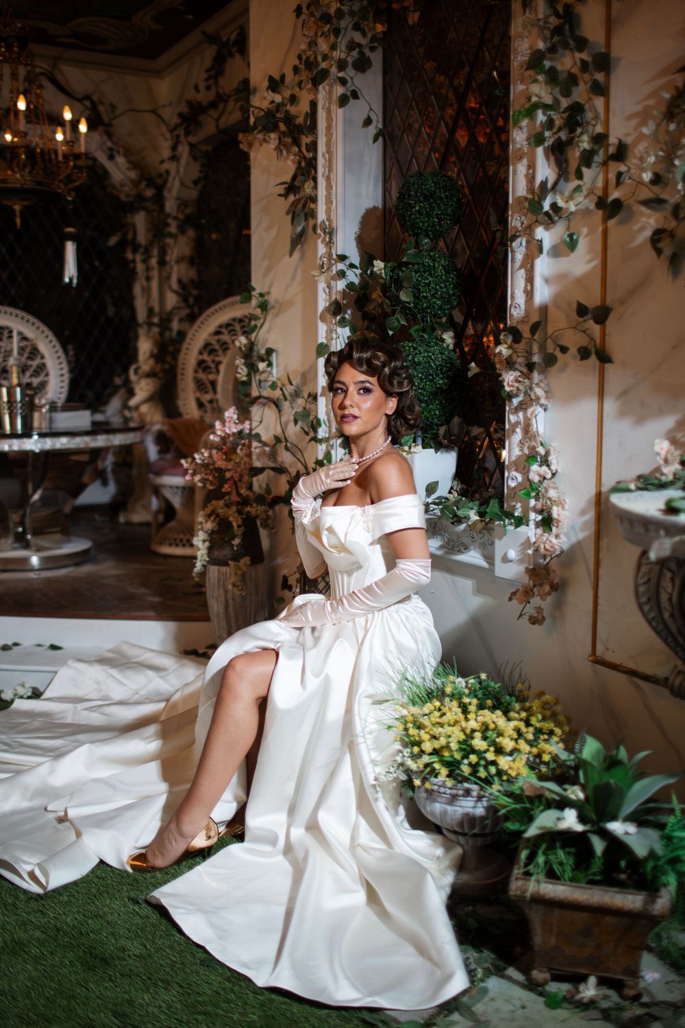 An elegant white off-shoulder wedding gown photographed in a romantic greenhouse setting with climbing vines and potted plants.