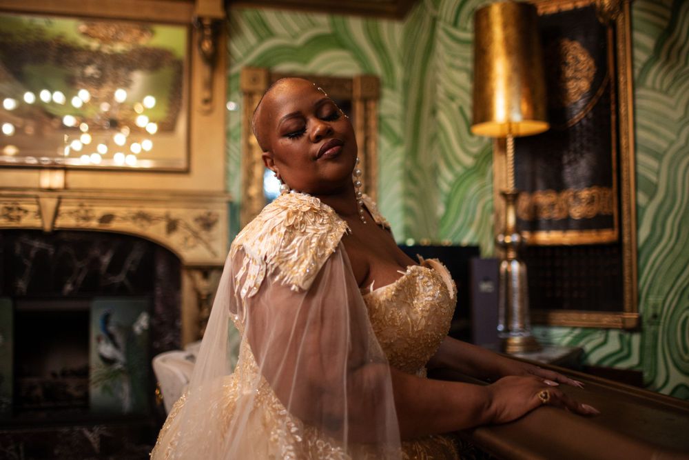 A person wearing a sheer, romantic vintage-style dress poses in an elegant room with palm wallpaper and antique decor.