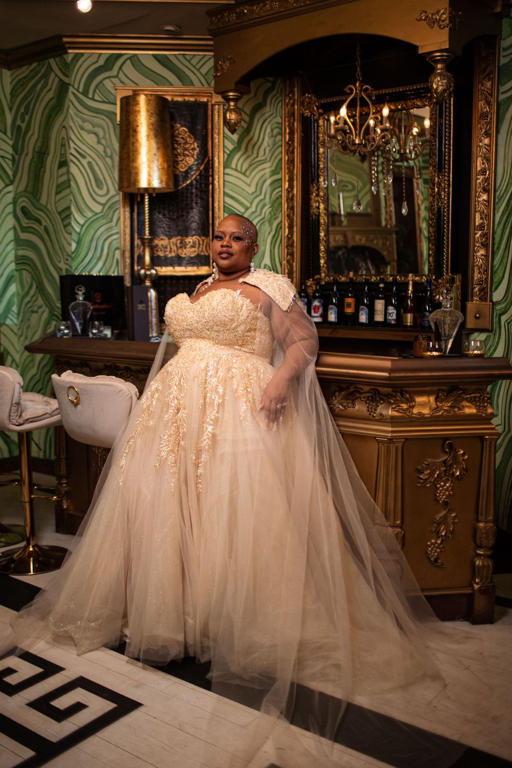 An elegant bridal gown with flowing tulle and lace detail is showcased in a luxurious bar setting with green marbled walls.