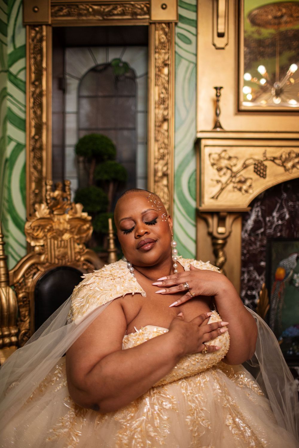 A bride in an elegant cream-colored lace gown poses dramatically on an ornate throne chair with gold detailing.
