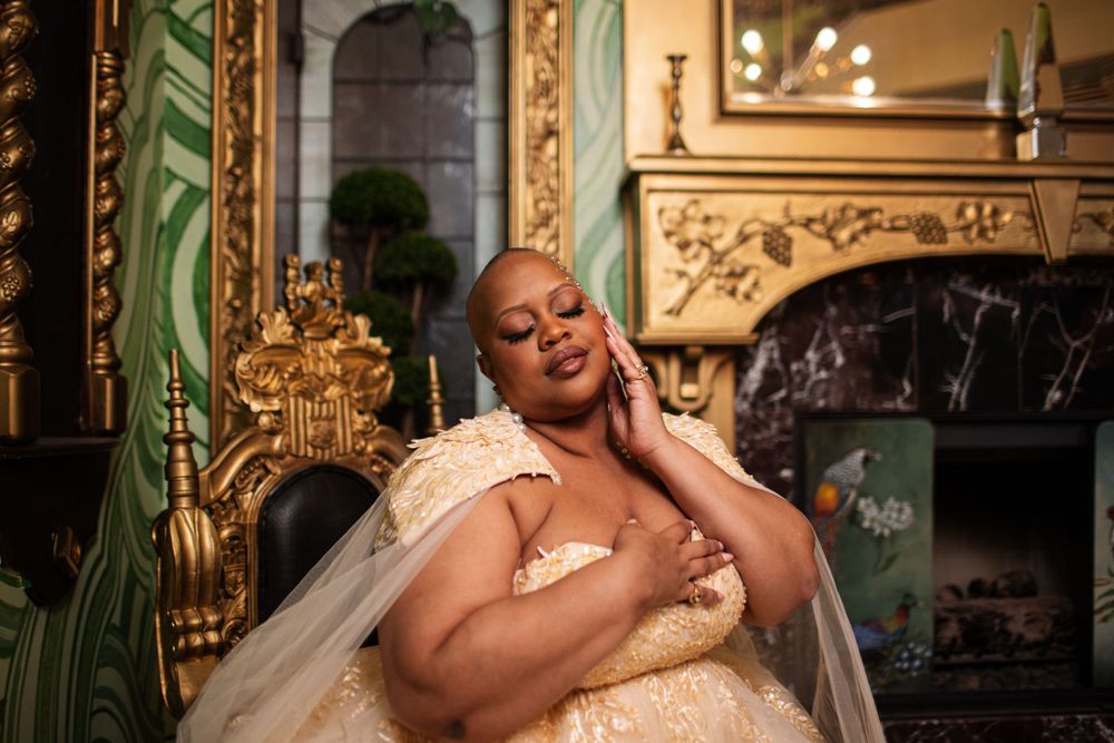 A bride in a cream wedding gown relaxes in an opulent room with ornate gold decor and a fireplace.