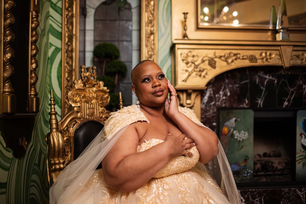 A bride in an elegant cream-colored gown poses thoughtfully in an ornate gold-trimmed interior setting.