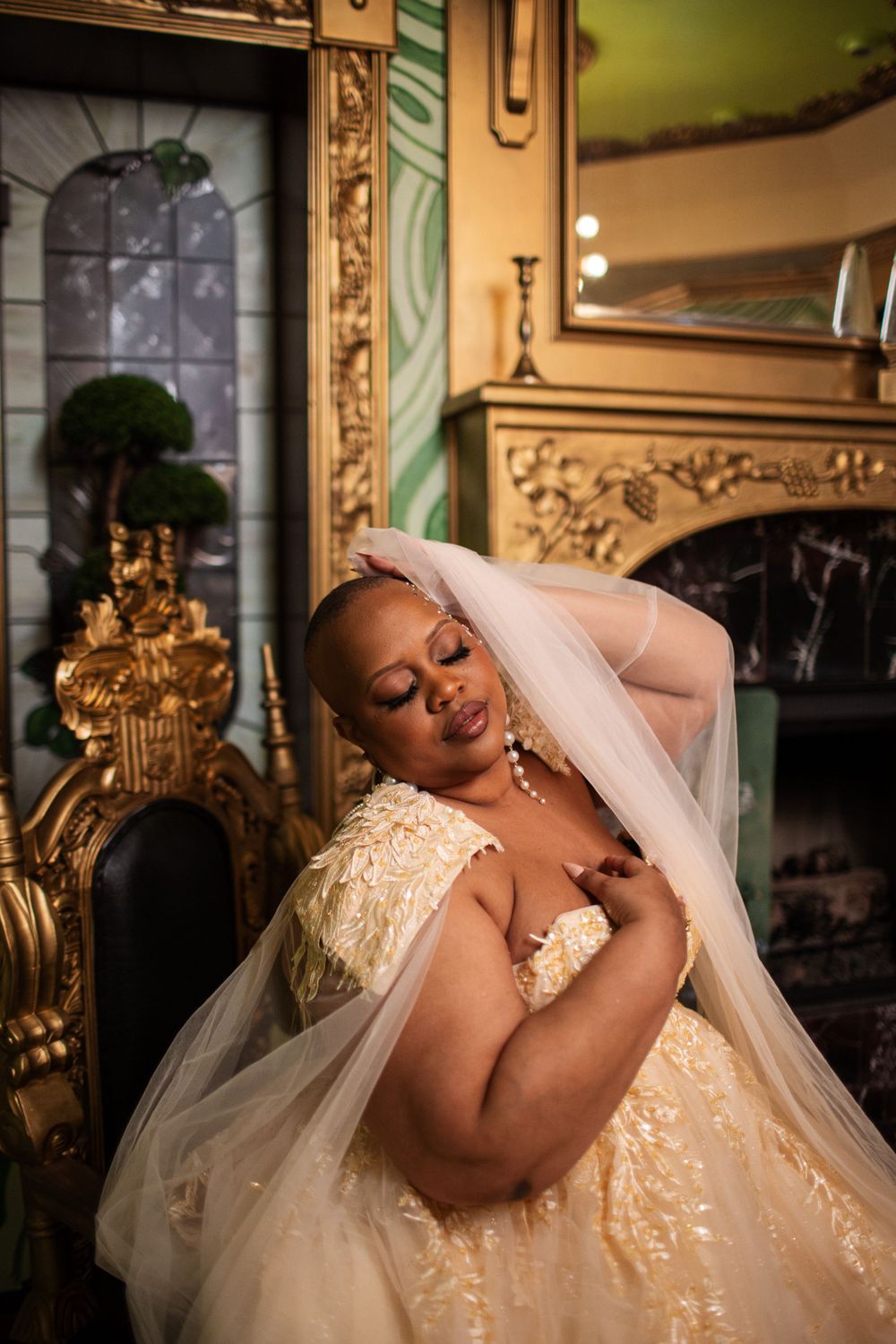 A bride in an elegant cream sequined wedding gown and veil poses in front of an ornate gold fireplace mantel.