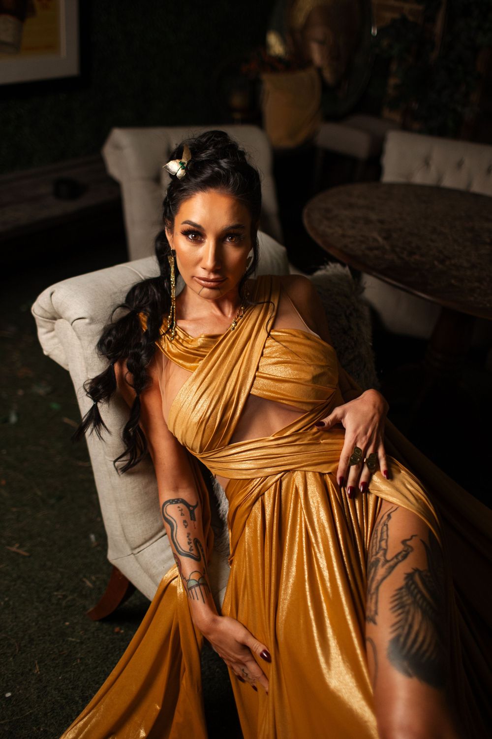 Woman in flowing golden dress with tattoos poses dramatically on white chair in moody lighting.