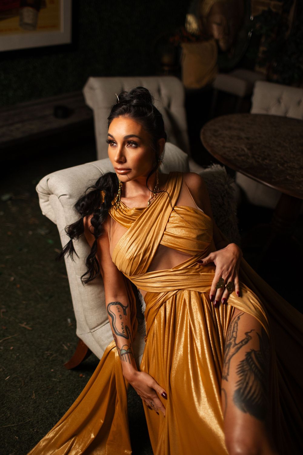 Model in draped gold dress with tattoos reclines on white armchair during glamorous photoshoot.