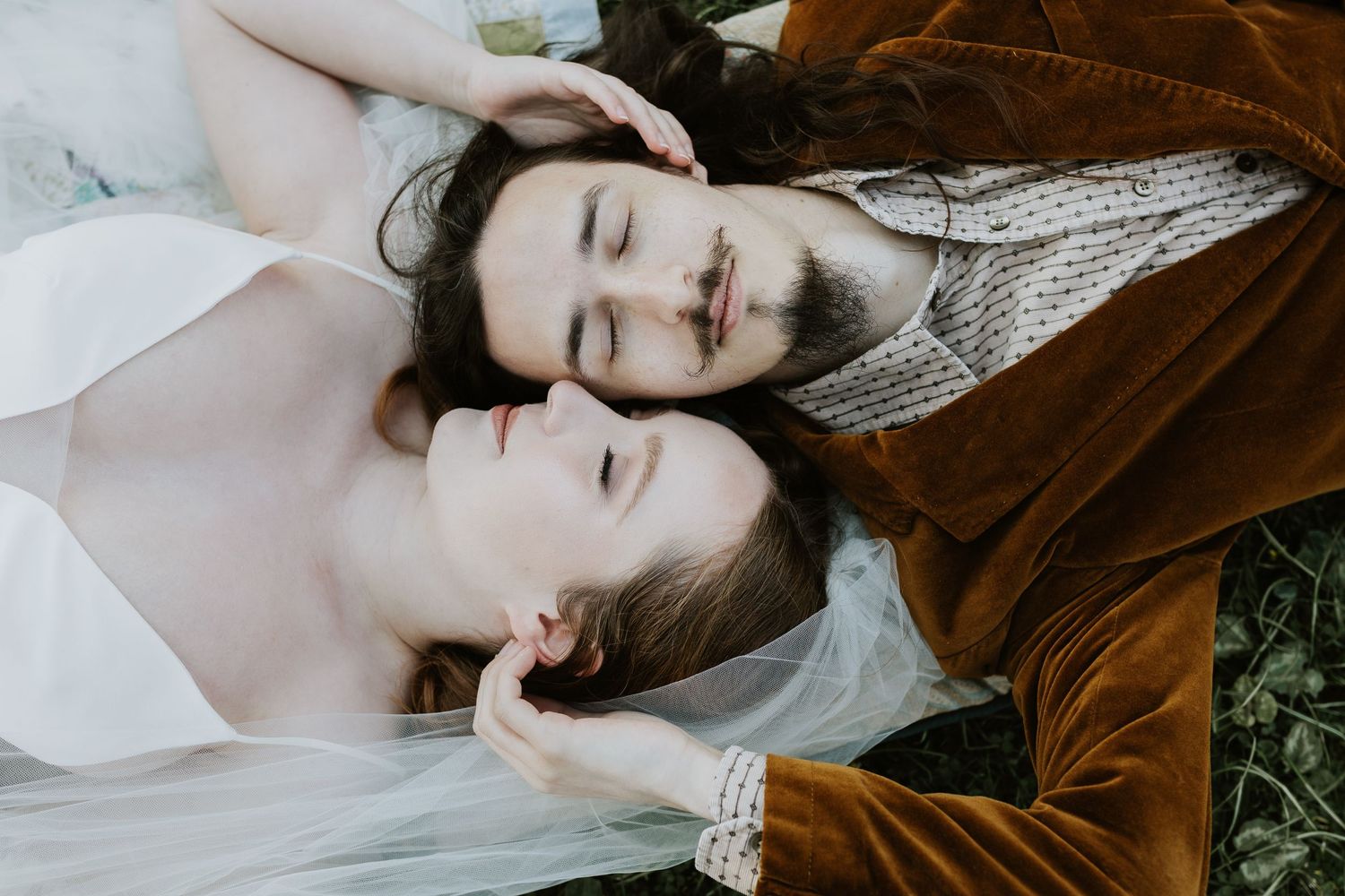 A couple laying on grass wearing white and brown velvet clothing, photographed from above in closeup.