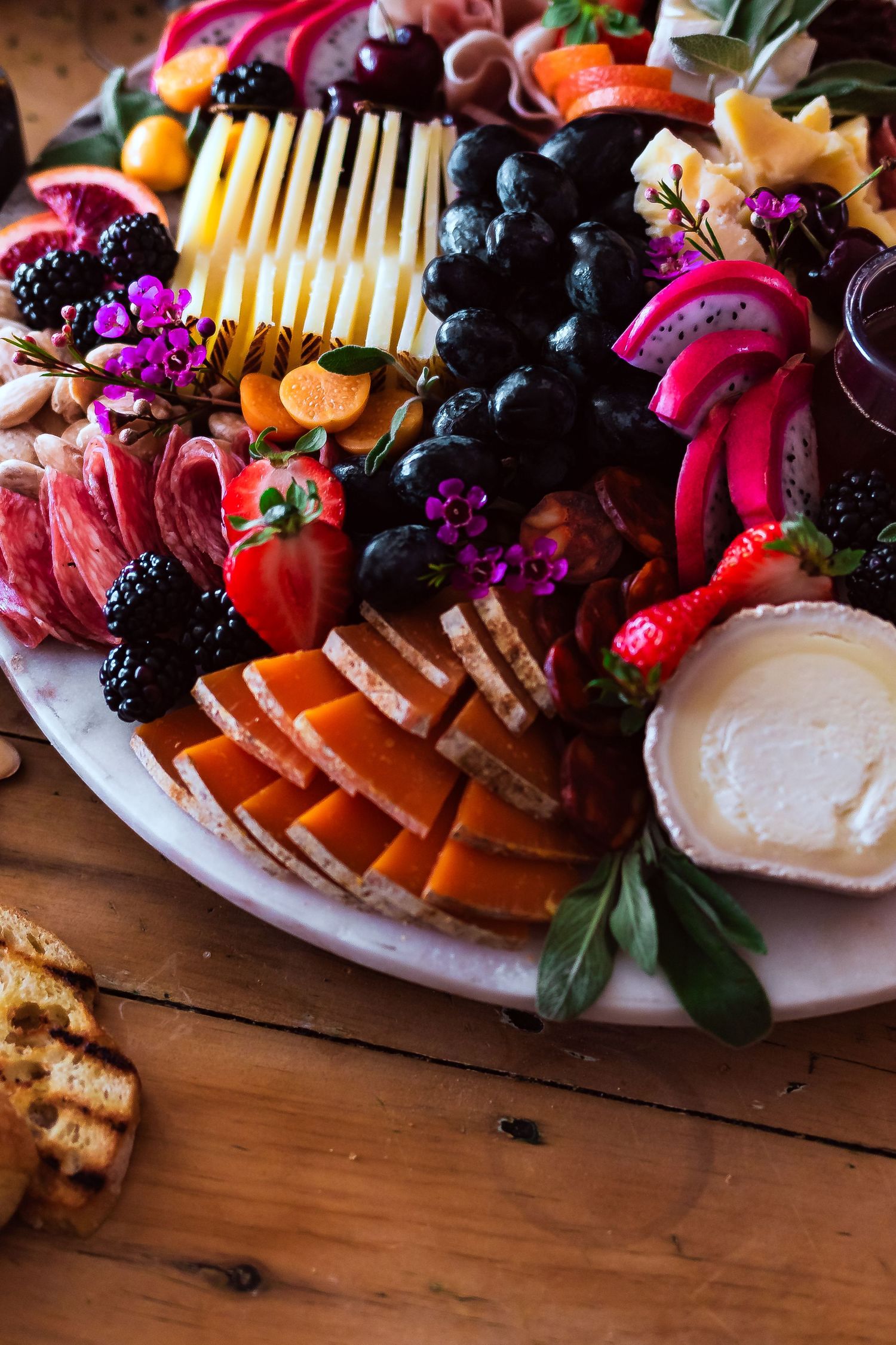 Artfully arranged charcuterie board with fresh grapes, cured meats, cheese slices, berries, and edible flowers on rustic wooden table.