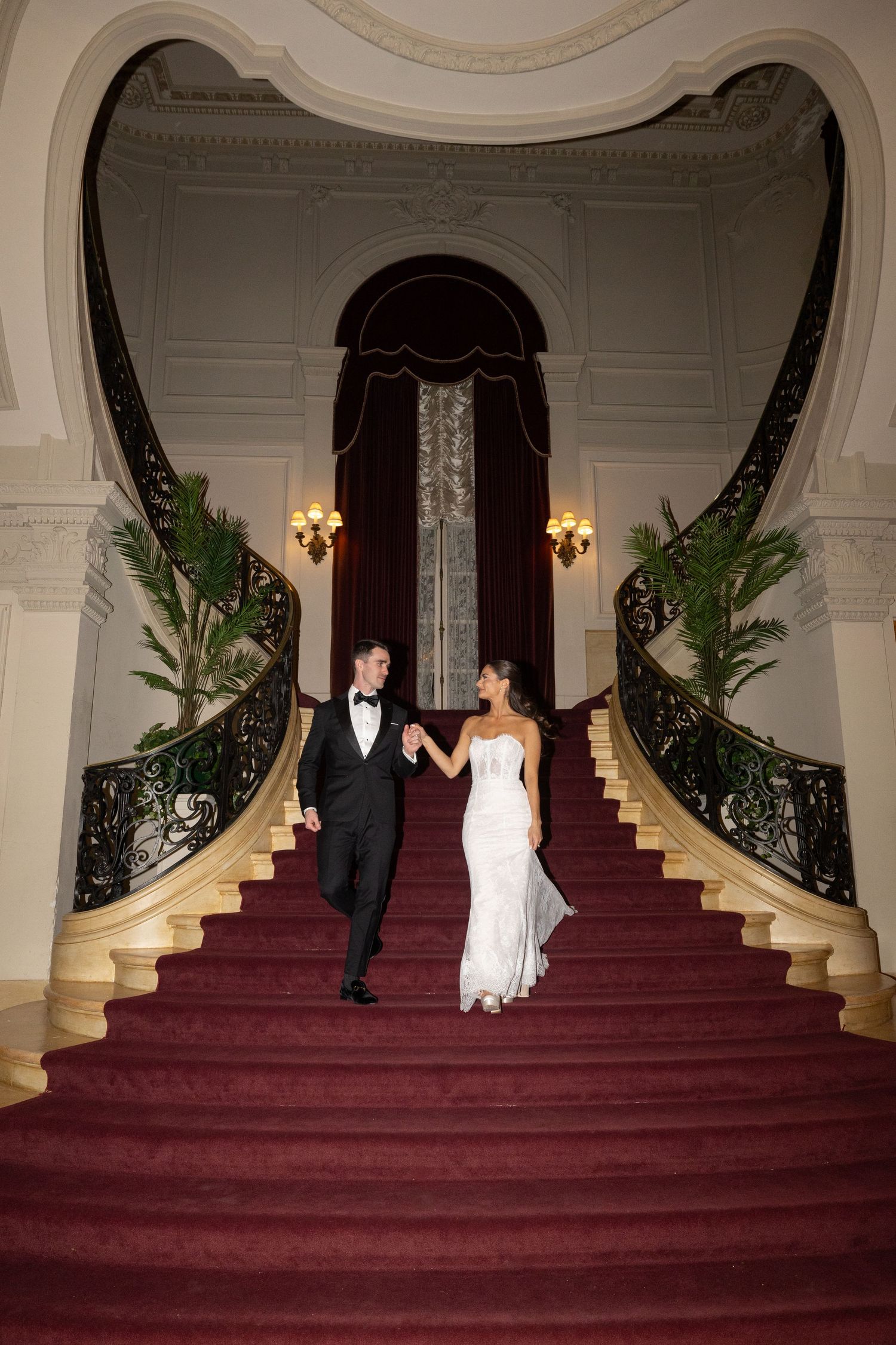Elegant couple walking down a grand staircase with red carpet and ornate railings in a luxurious venue.