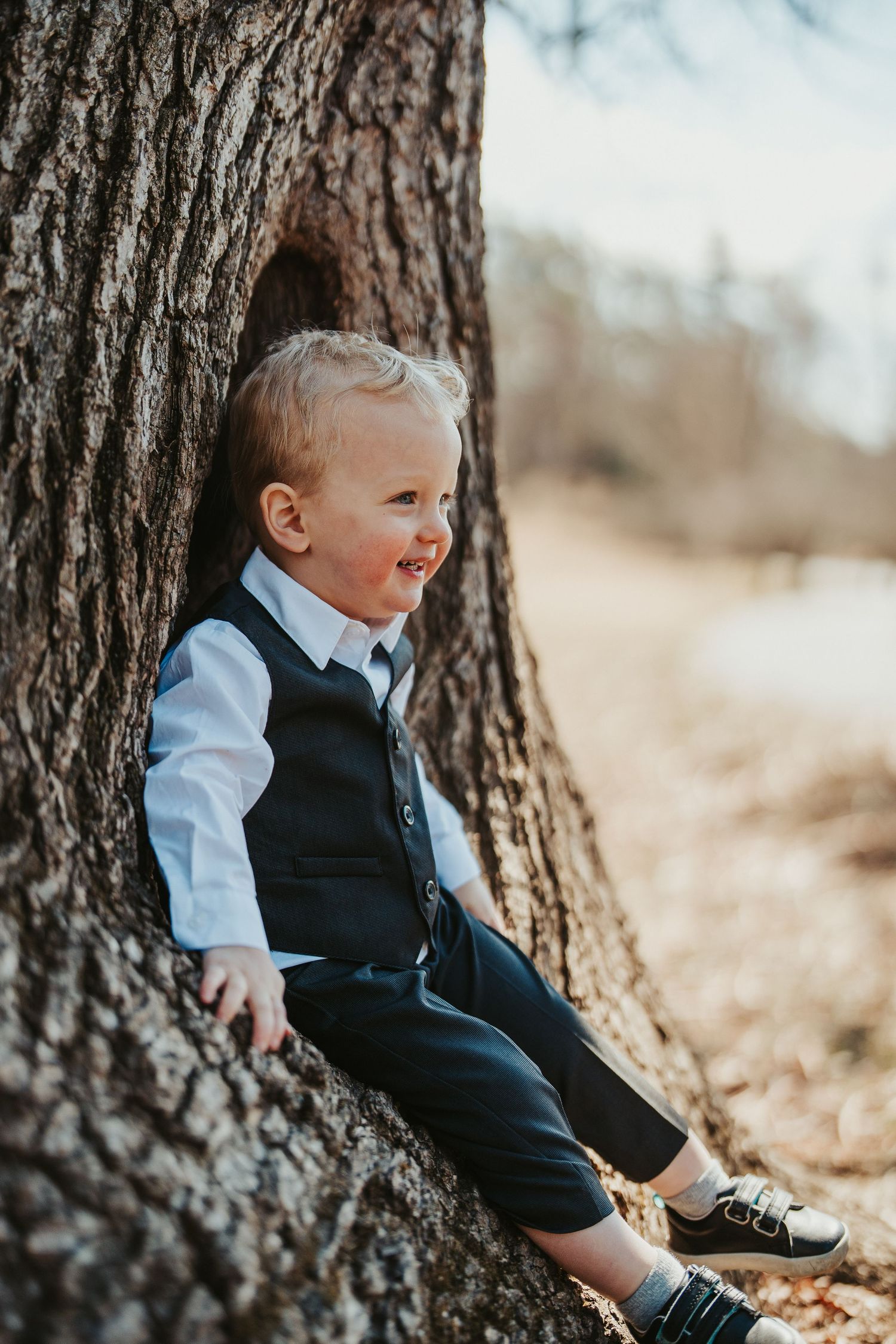 A young child in formal attire laughs joyfully while sitting in a hollow of a large tree trunk in autumn.