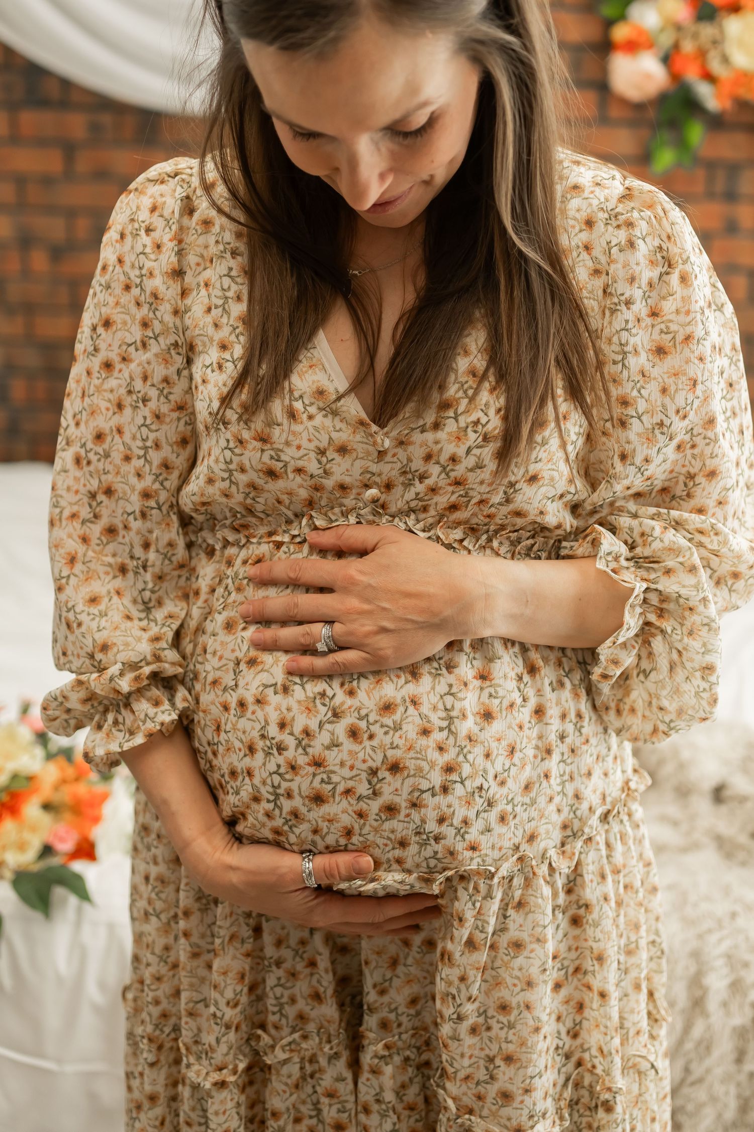 Person wearing floral dress tenderly holding pregnant belly in front of brick wall with white drapes.