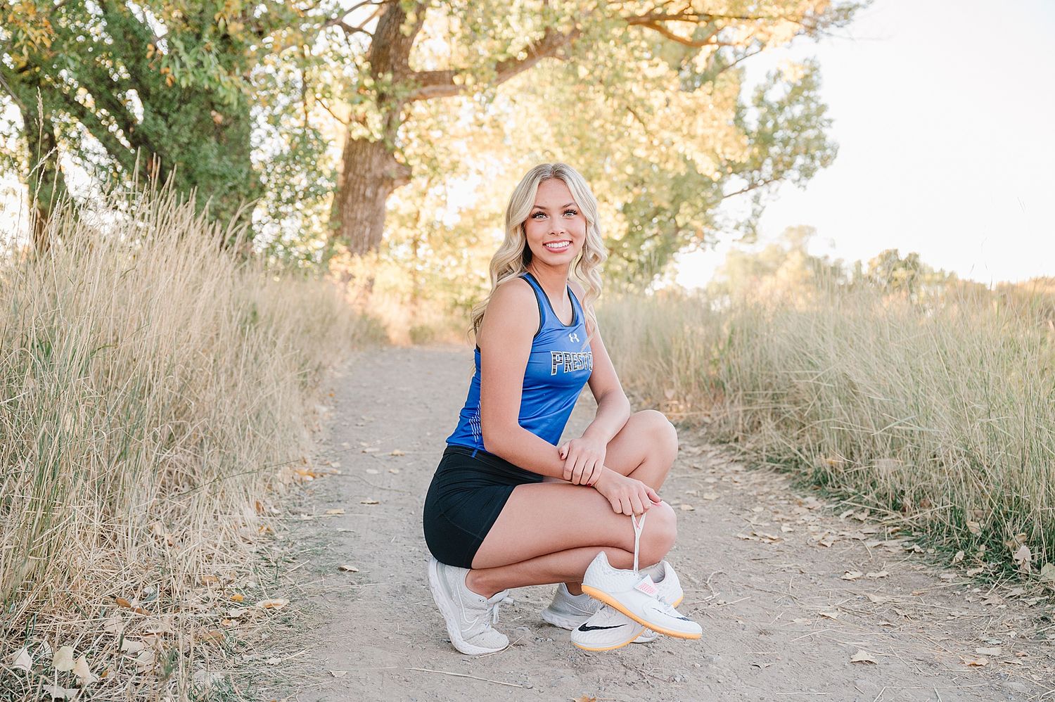 Athletic person in blue tank top and black shorts crouches on a dirt path surrounded by tall grass during golden hour.