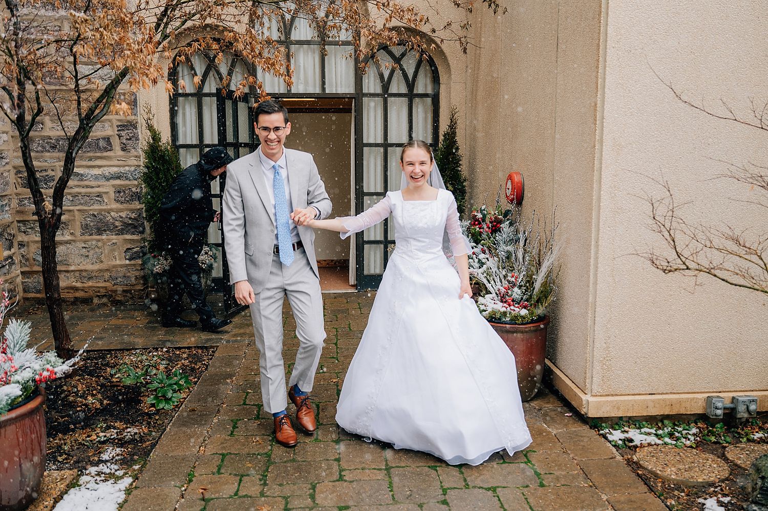 A couple in wedding attire poses together on a stone walkway outside a church with gothic windows and autumn foliage.