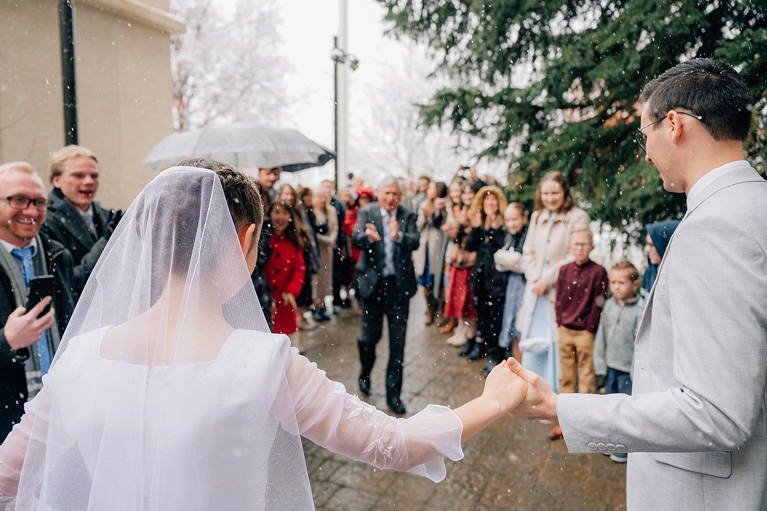 A couple celebrates during their winter wedding ceremony as guests look on with umbrellas in the misty weather.
