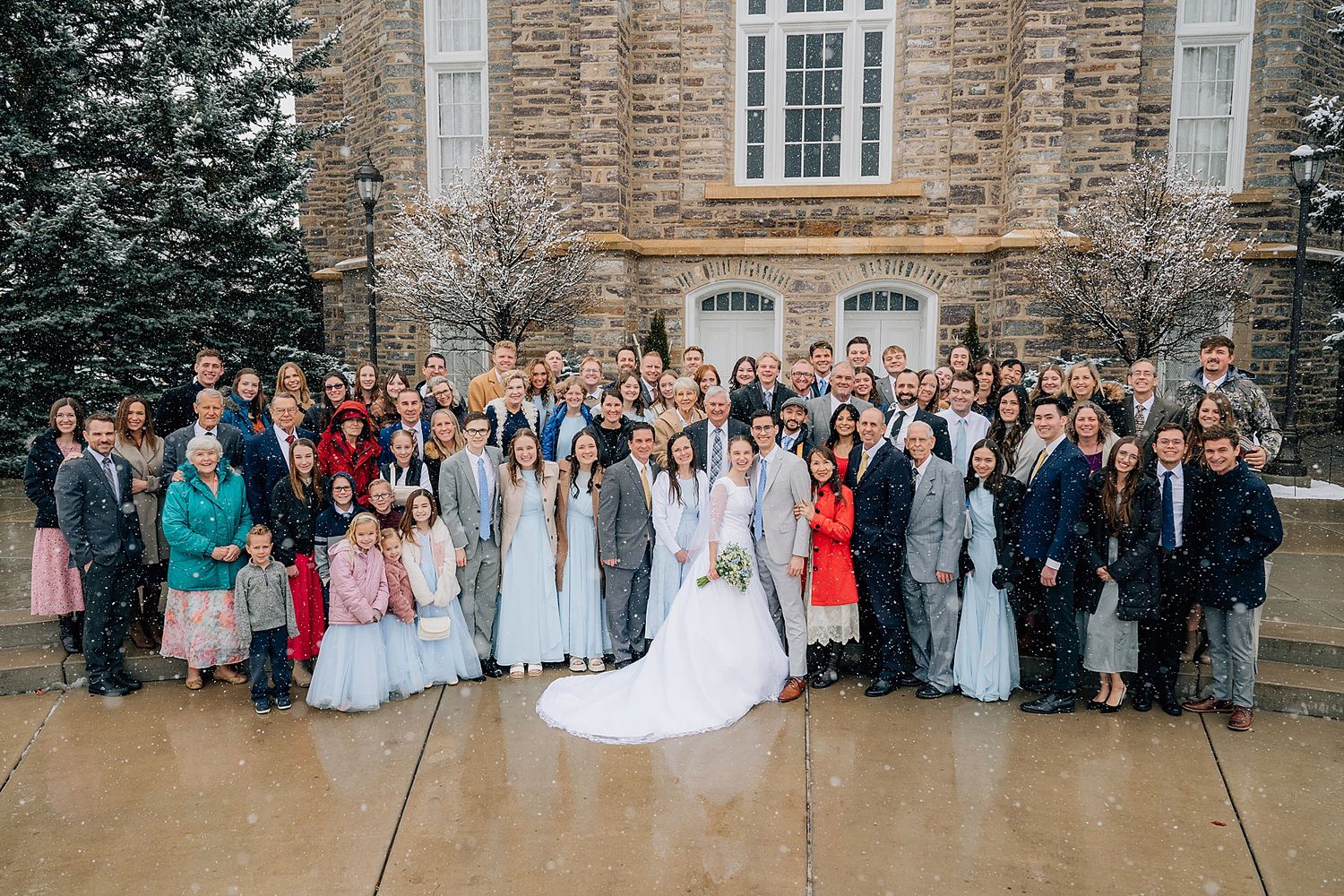 Large wedding party gathers for group photo outside stone building on overcast day.
