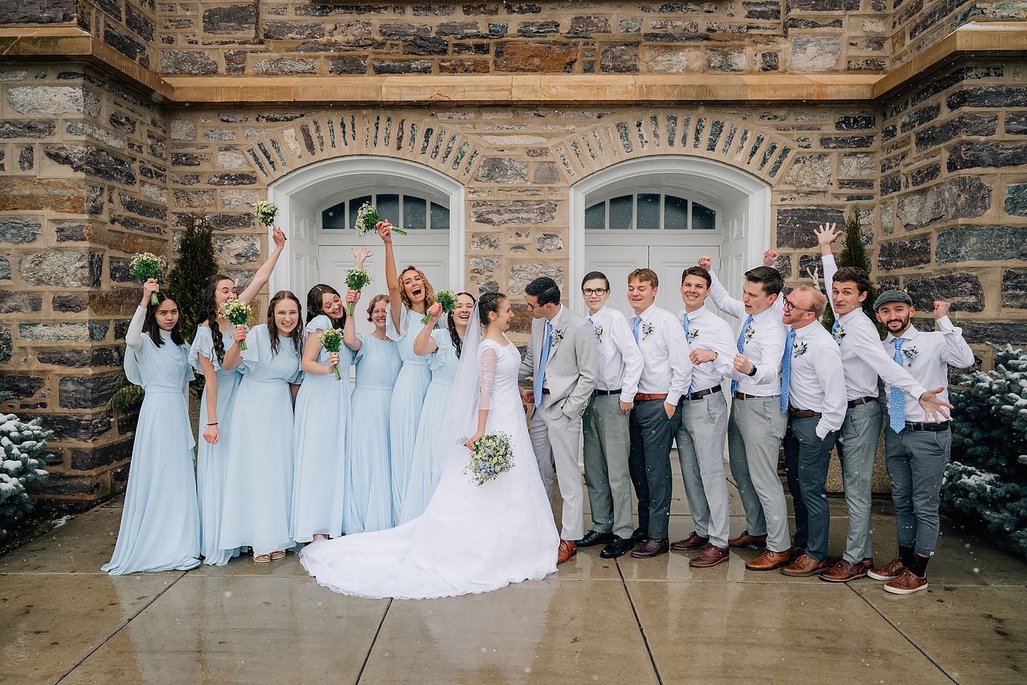Wedding party in light blue and gray attire pose together outside stone building with arched windows.